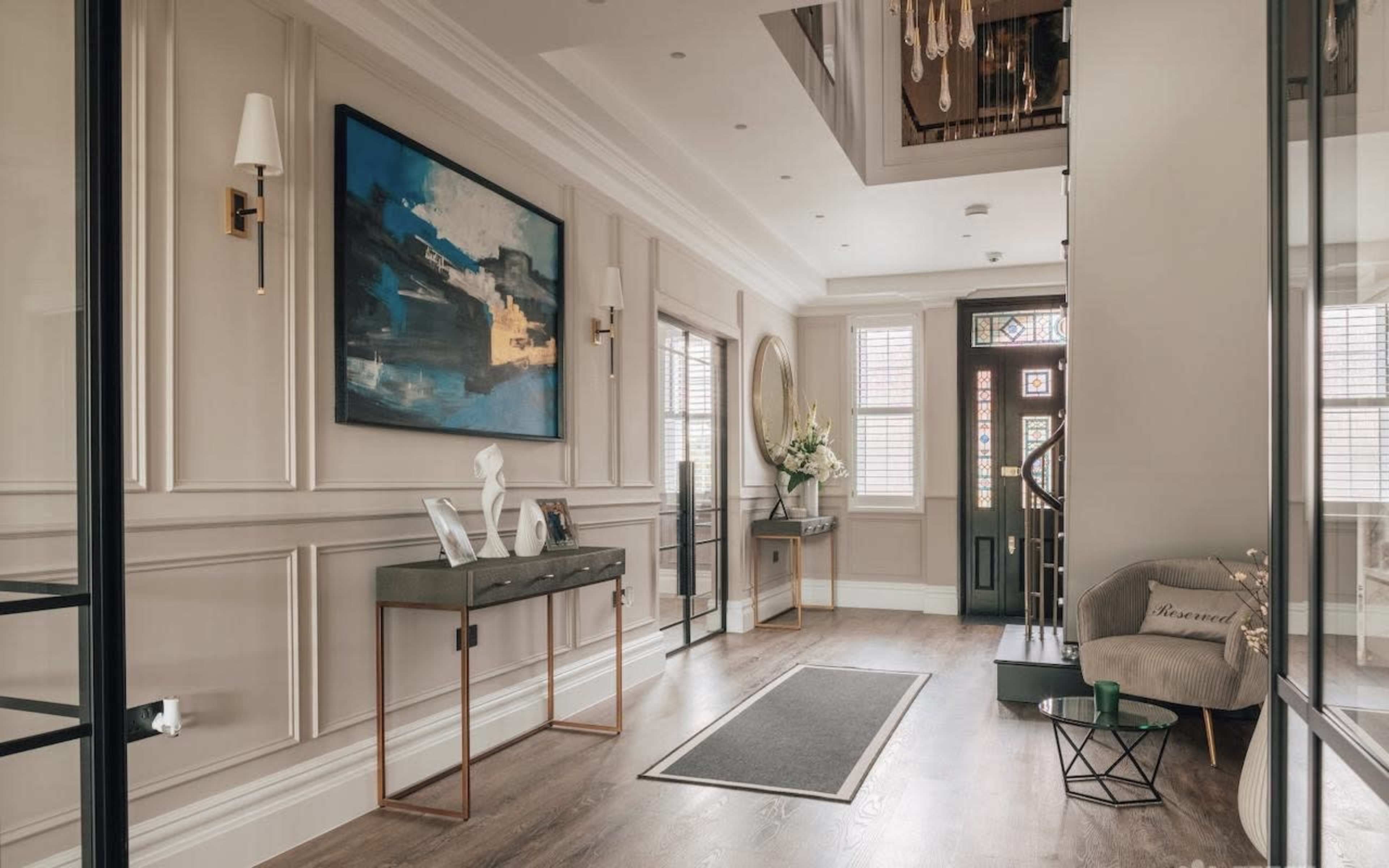 The image shows a spacious hallway with a console table, a framed abstract painting, a mirror, and a staircase leading to an upper level, illuminated by natural light.