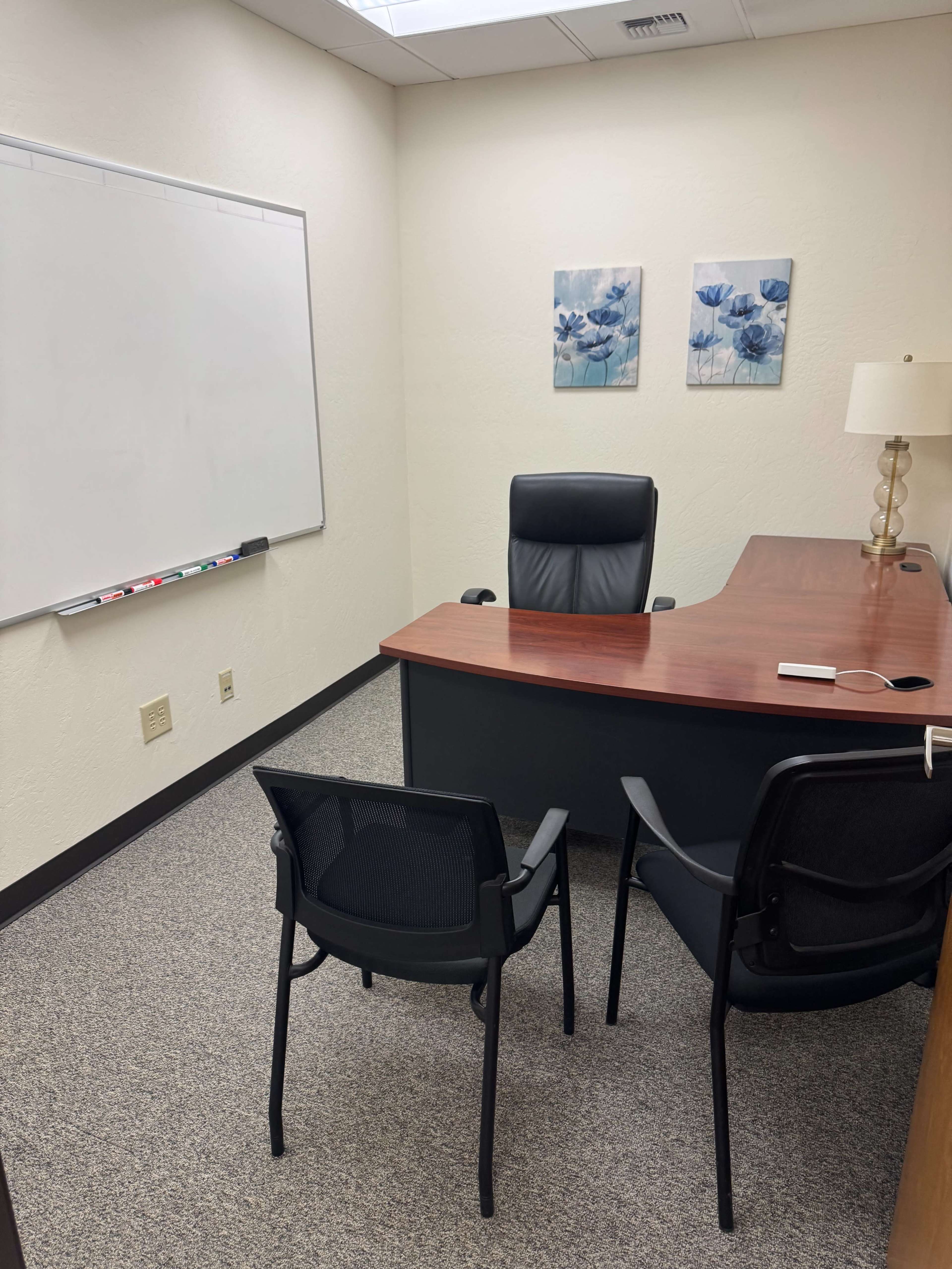 The image shows a tidy office with a desk, three chairs, a whiteboard, and two framed floral artworks on the wall.