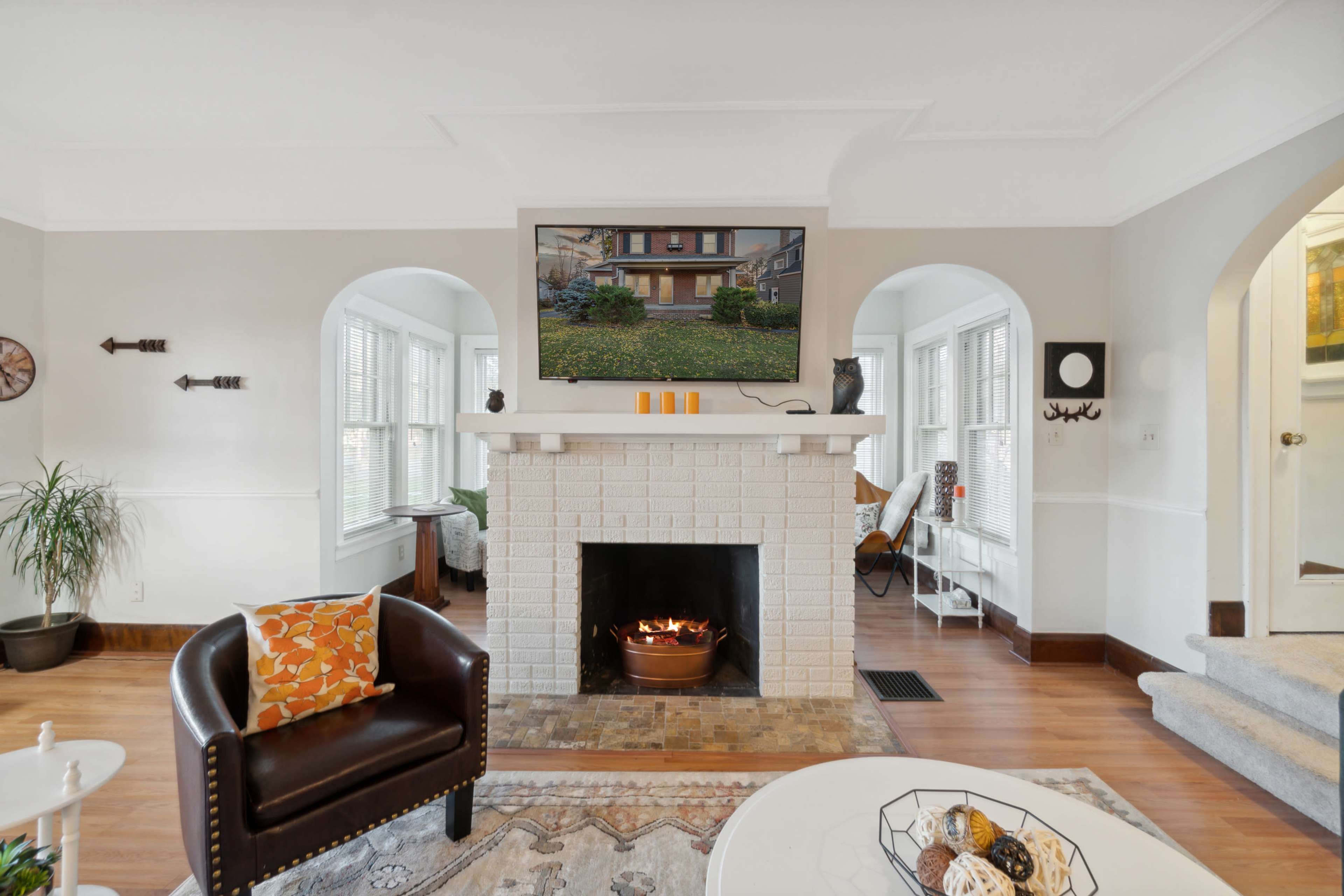 The image depicts a cozy living room featuring a white brick fireplace with a mounted screen above, a brown leather armchair, and large windows allowing natural light to fill the space.