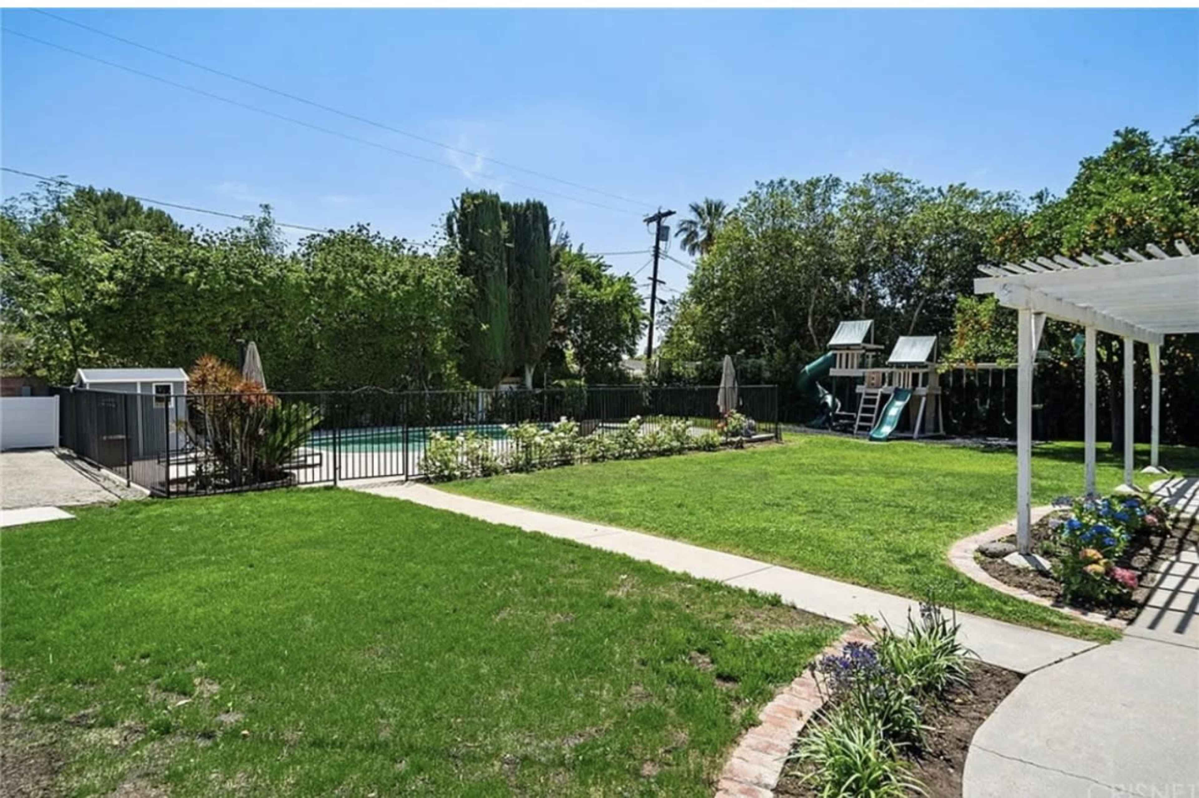 The image shows a landscaped backyard with a green lawn, a fenced swimming pool, and a playset in the corner.