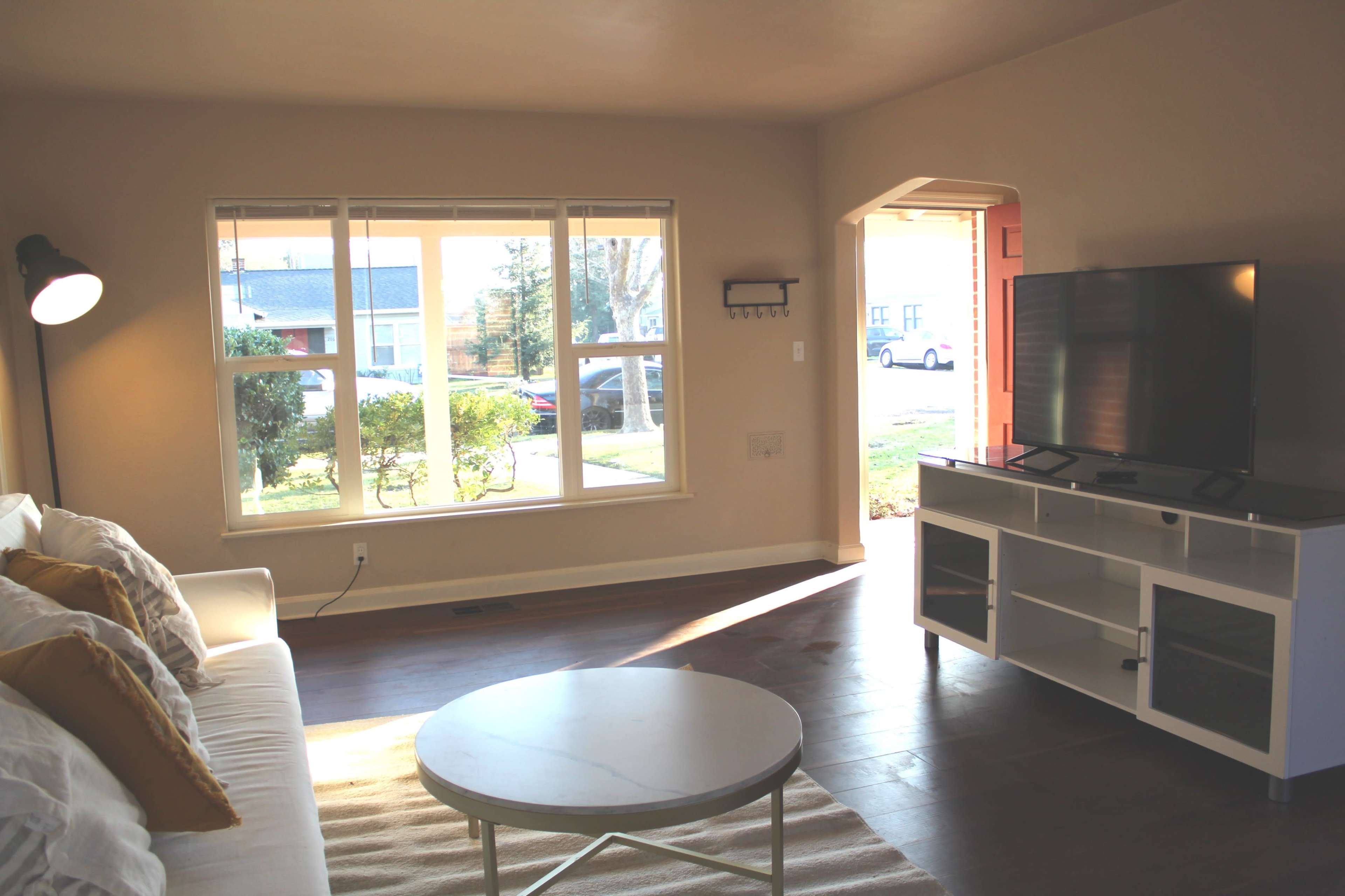 The image shows a light-filled living room with a white sofa, a round coffee table, and a television unit, featuring a view of a front yard through large windows.