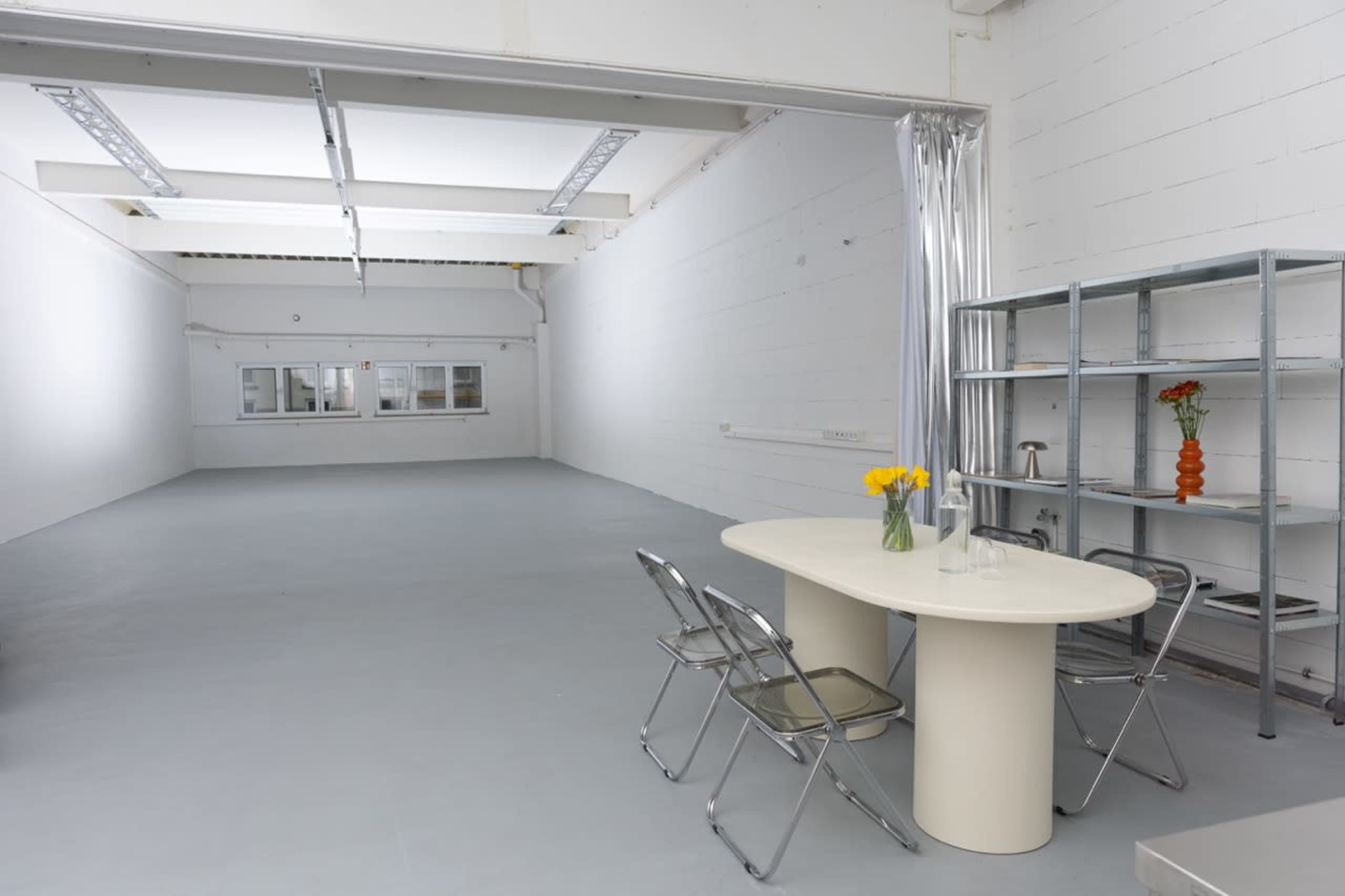The image shows a minimalistic, empty room with white walls and a round table set with a vase of flowers near a shelf.