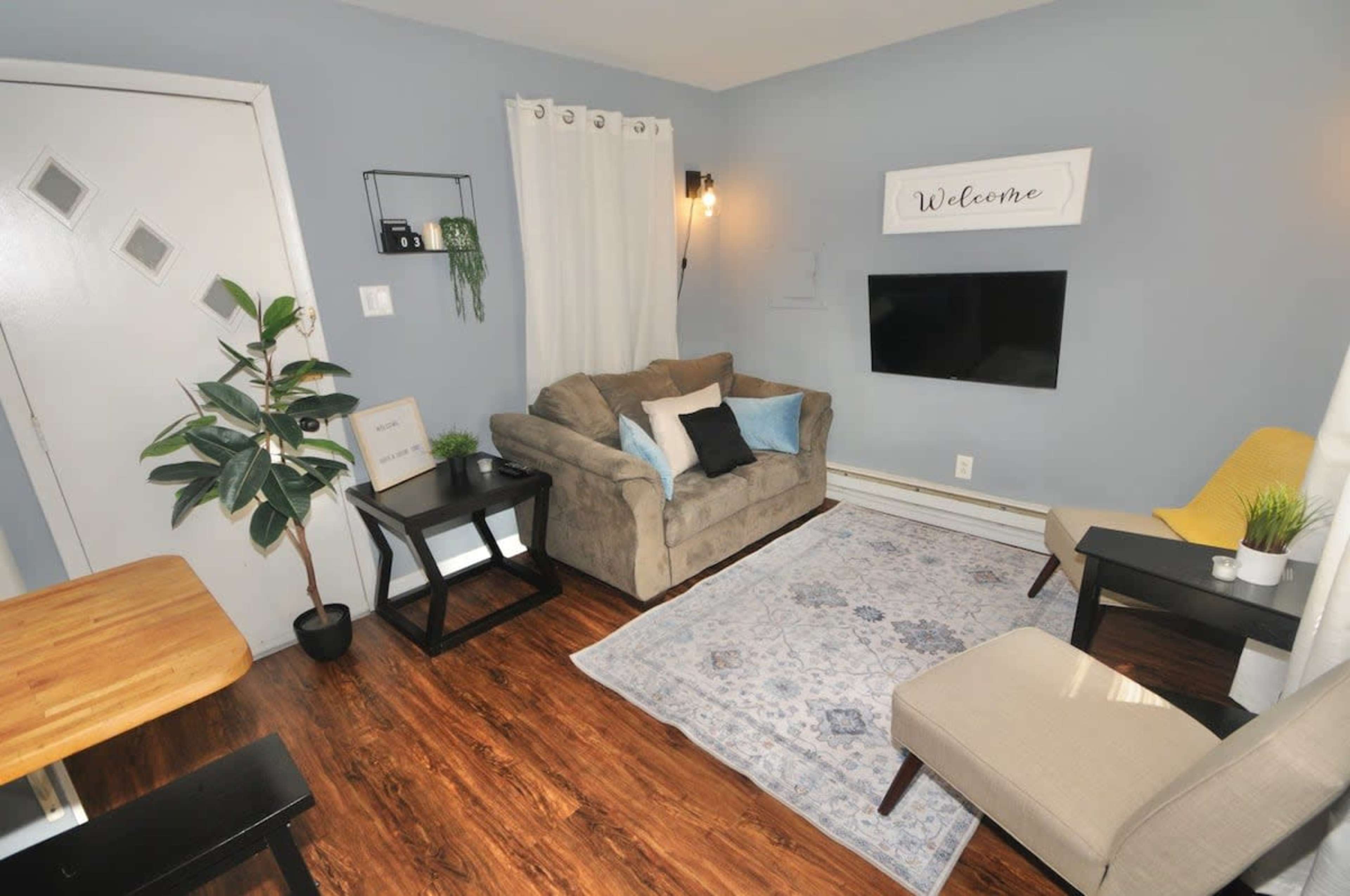 A cozy living room features a couch, two chairs, a coffee table, a TV mounted on the wall, and decorative plants against light blue walls.