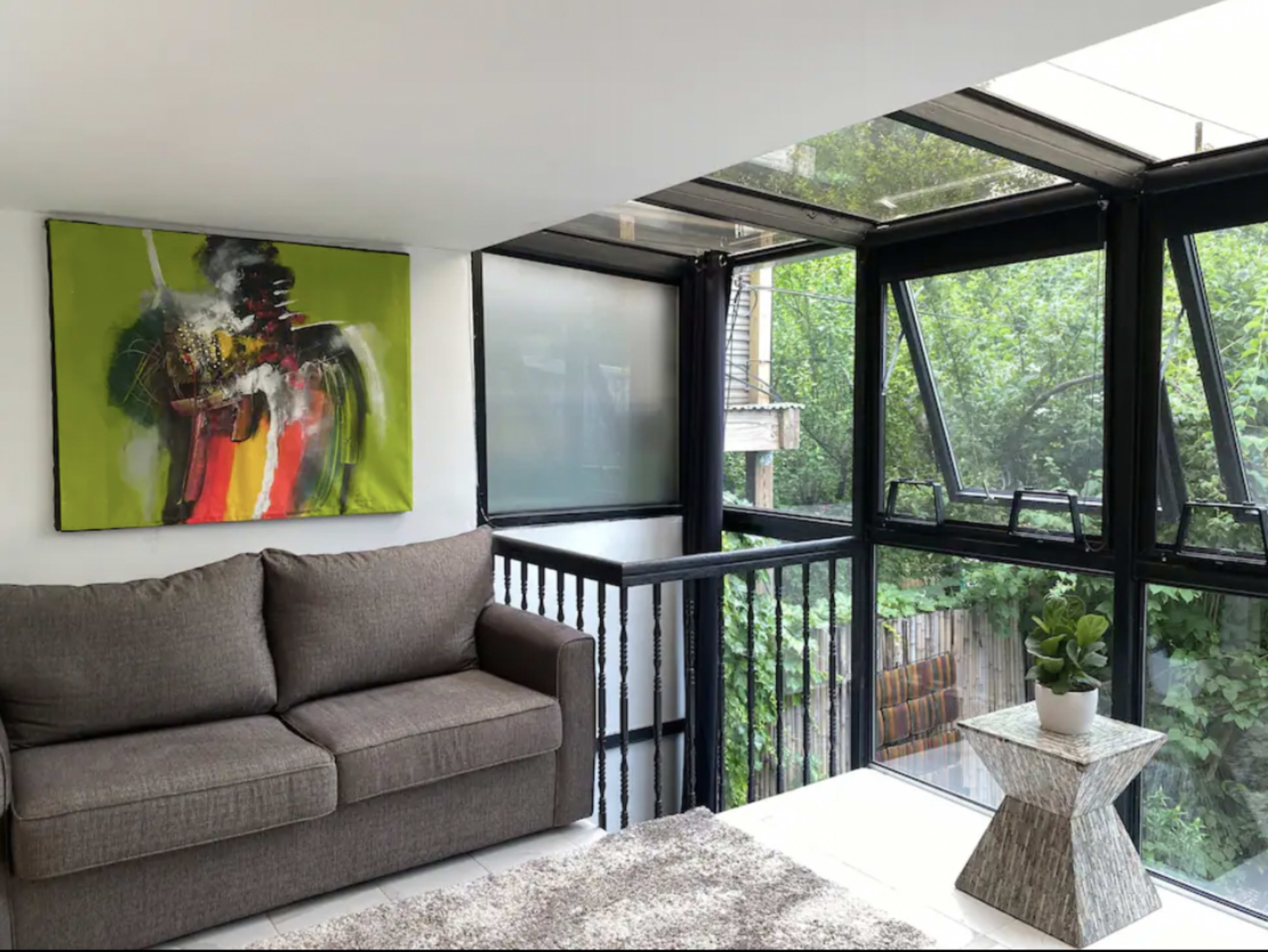 The interior features a gray sofa facing a large window, accompanied by a modern coffee table and a colorful abstract painting on the wall.
