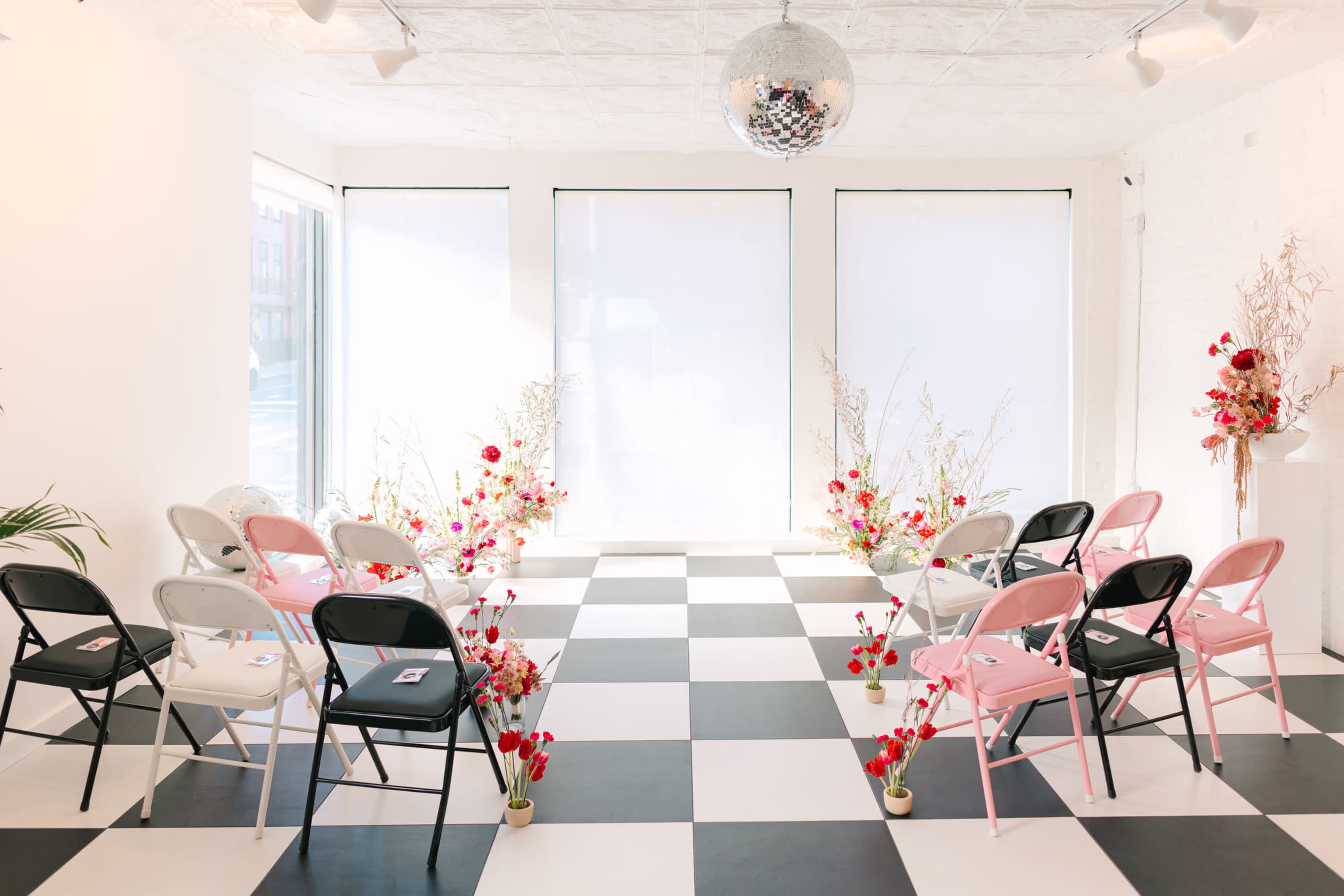 A modern indoor event space features a checkerboard floor with black and white chairs arranged around floral decorations.