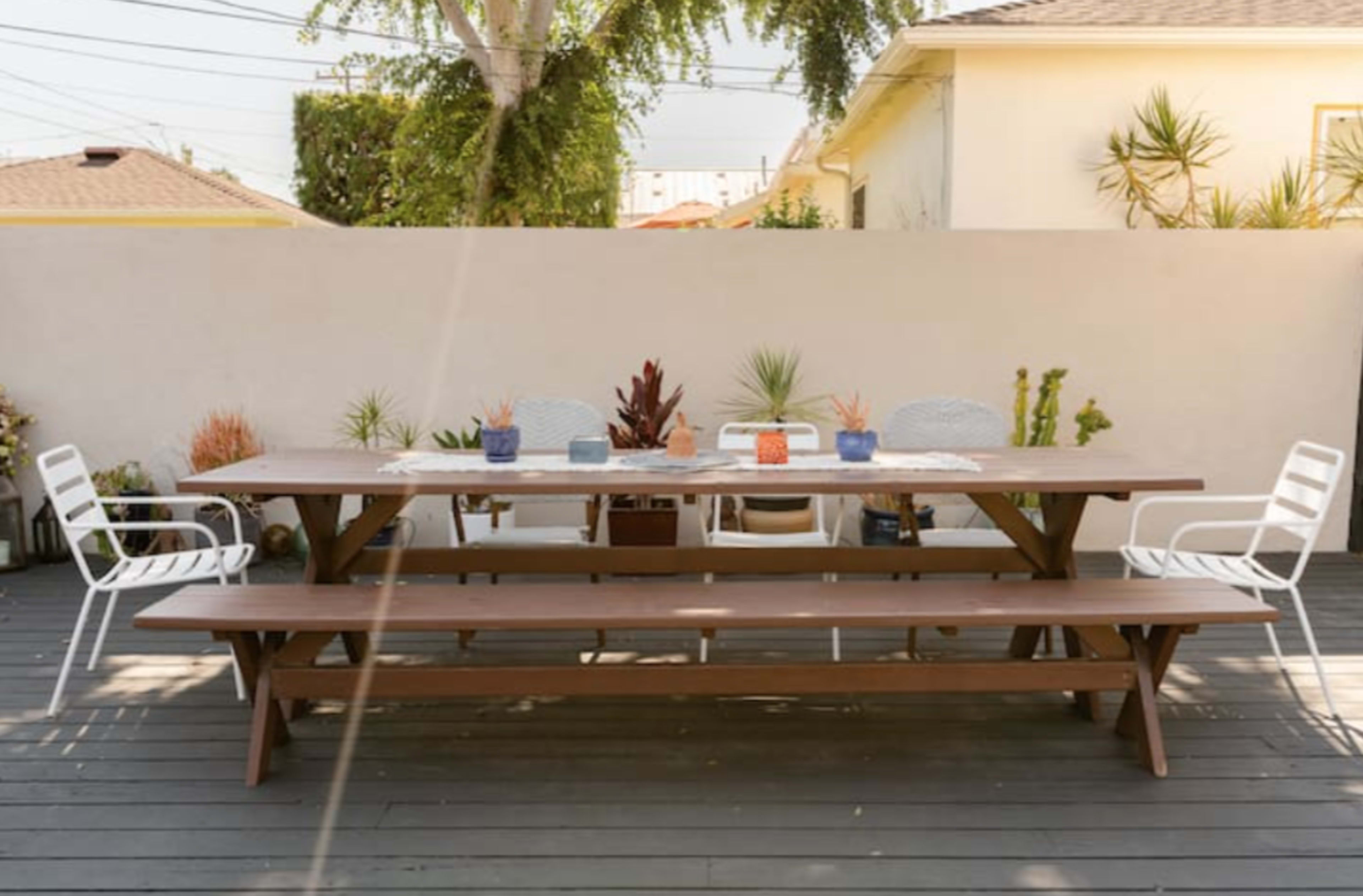 A spacious wooden dining table set in a minimalistic outdoor area with potted plants and two white chairs on either side.