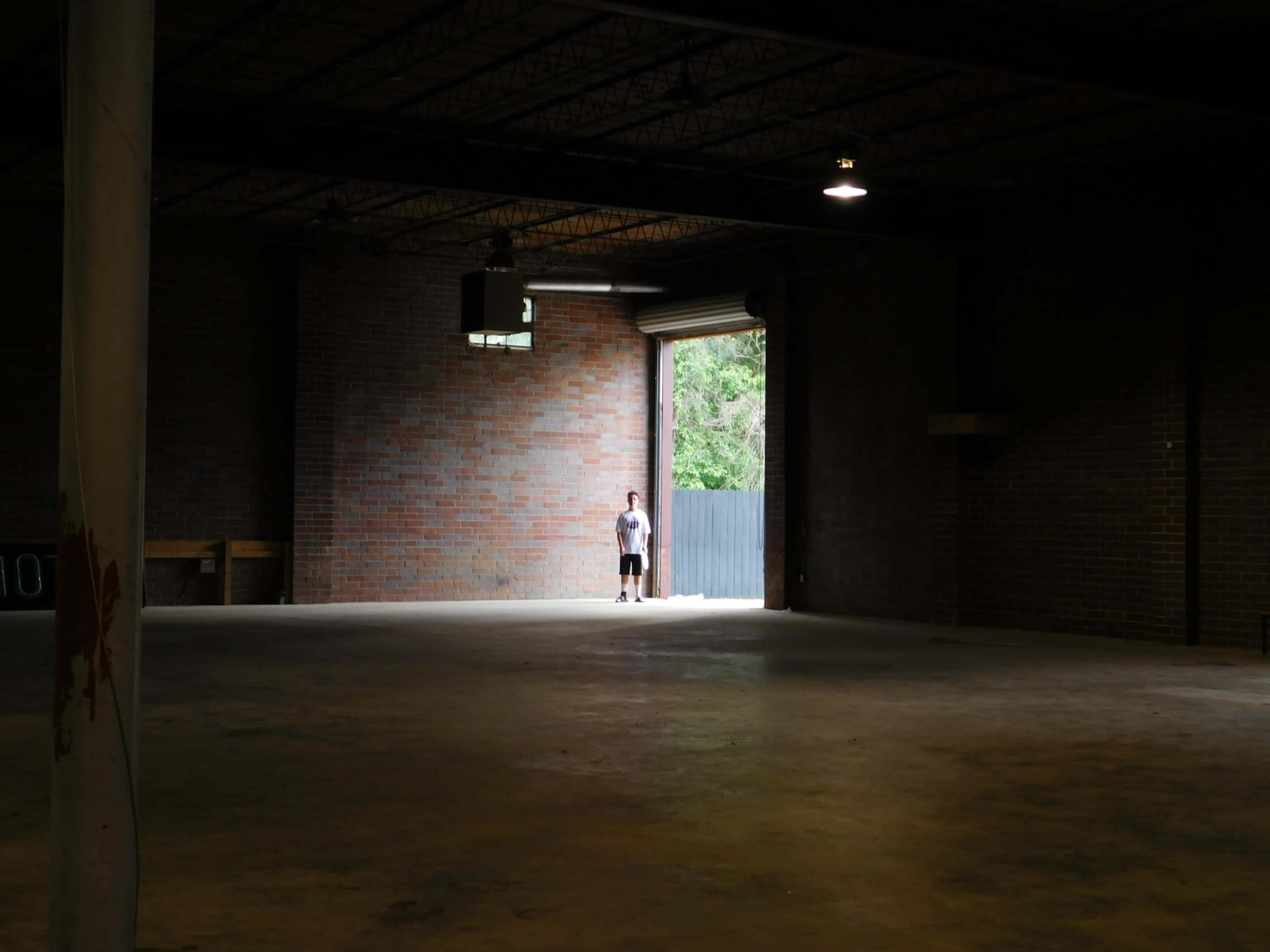 A person stands in an empty warehouse with a large open door at the far end, allowing natural light to flood the space.