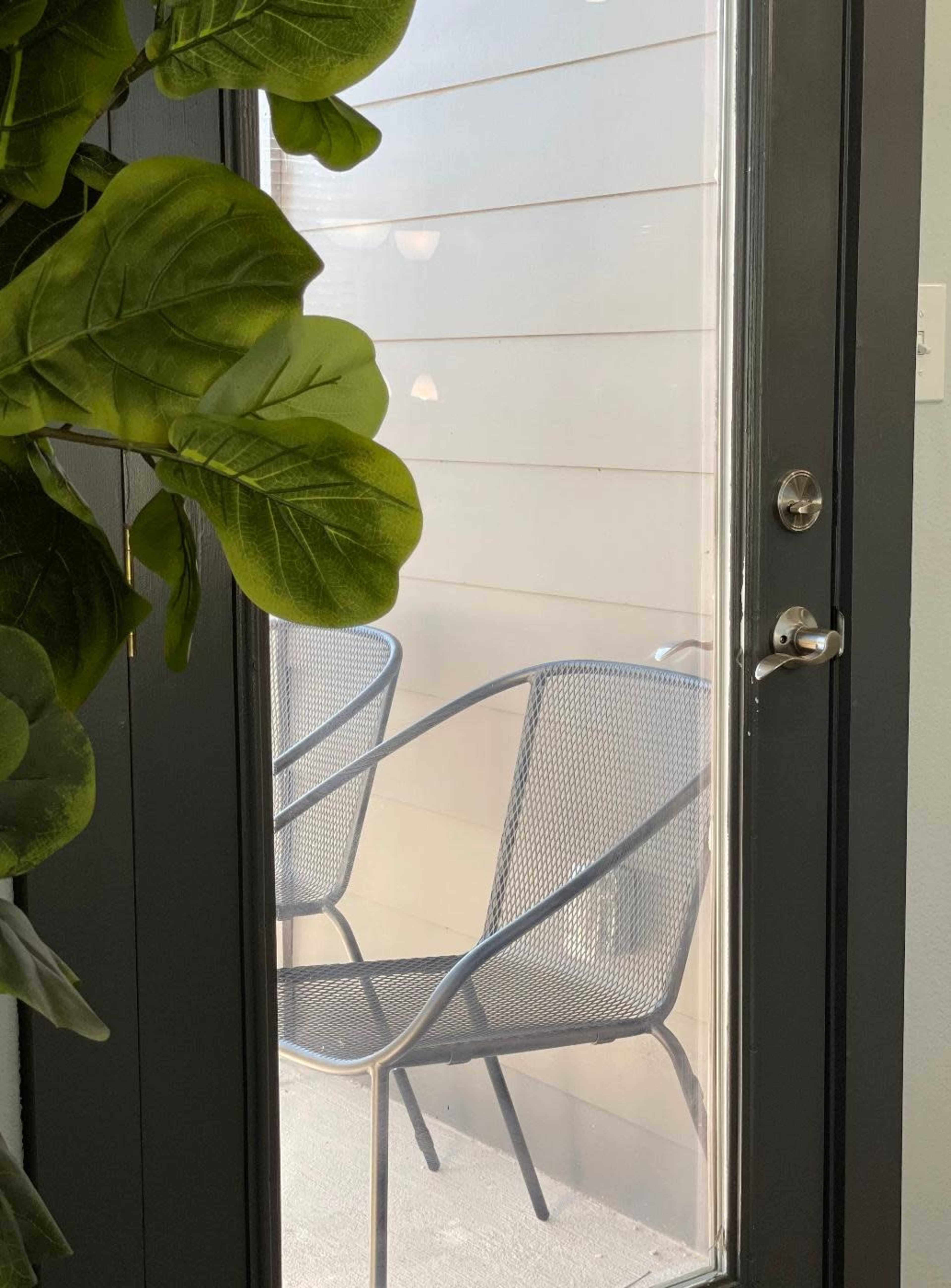 A glass door frames two metal chairs on a balcony, with a large green plant partially obscuring the view.
