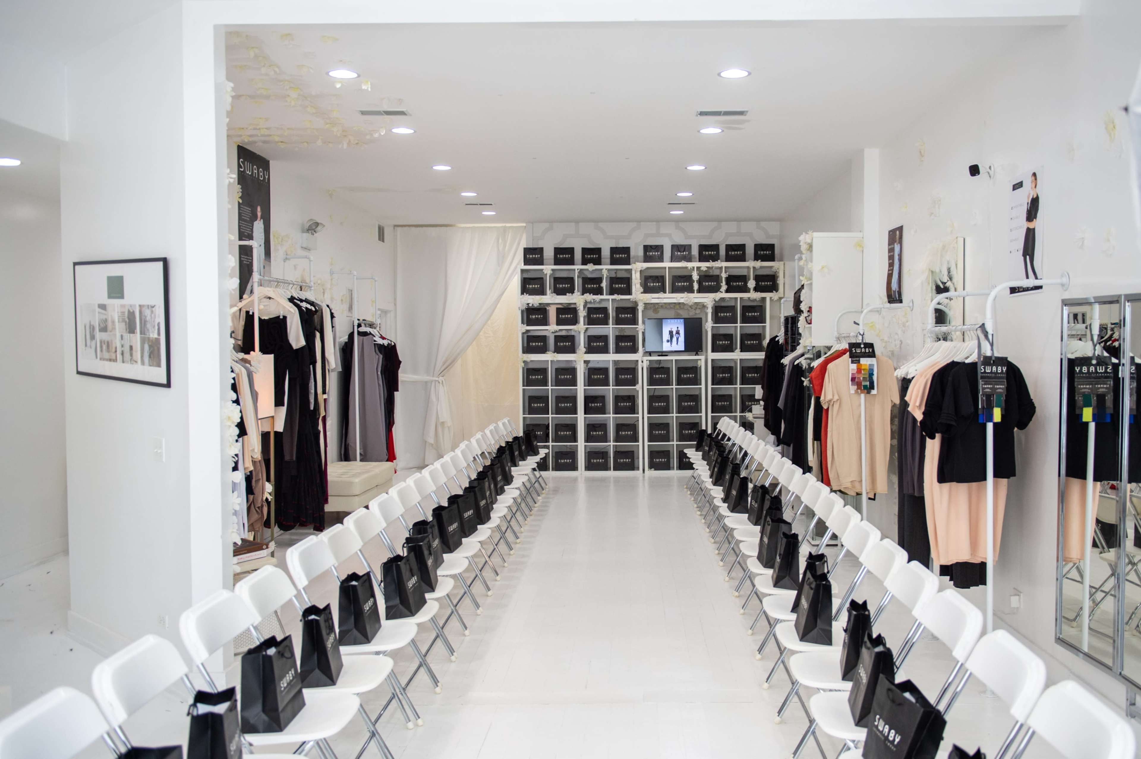 A fashion showroom with two rows of white chairs facing a presentation area flanked by clothing racks and storage units.