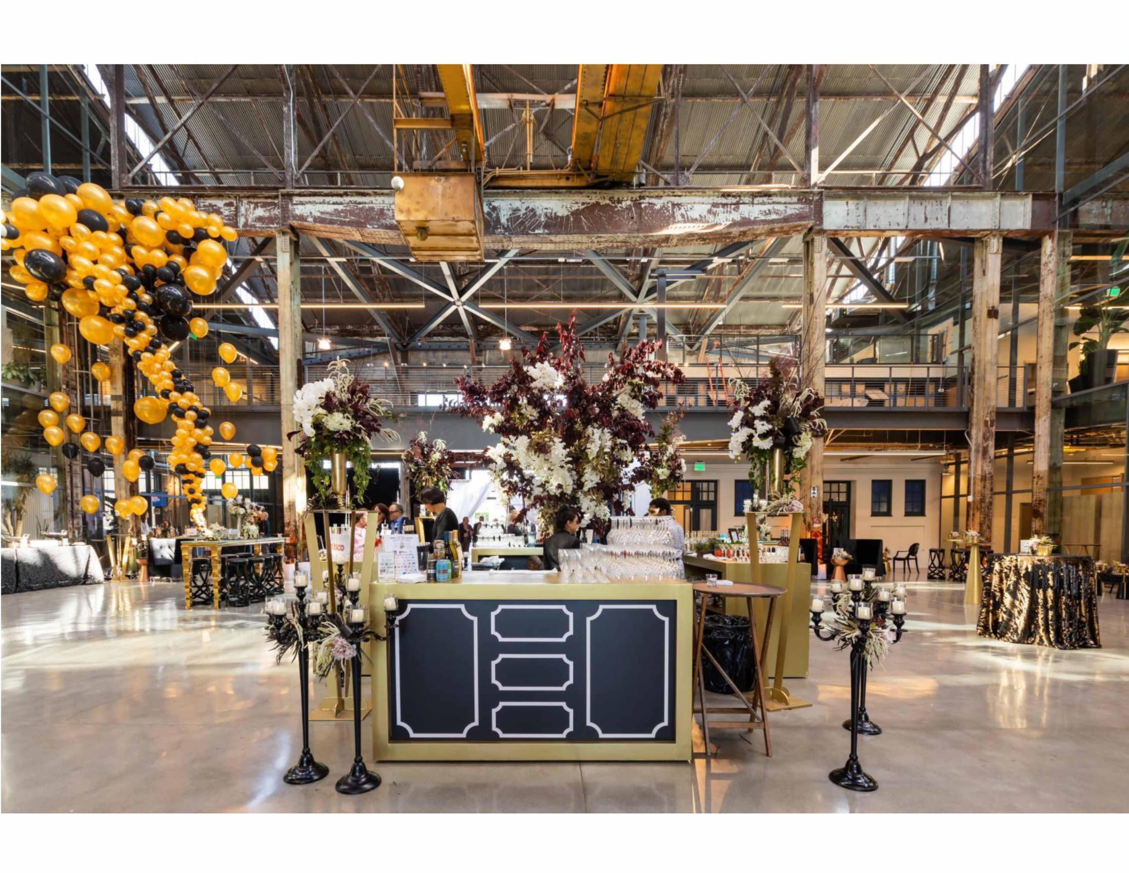A large industrial space is set up for an event, featuring a central bar adorned with flowers, surrounded by tables and decorations in black and gold, with balloons floating above.