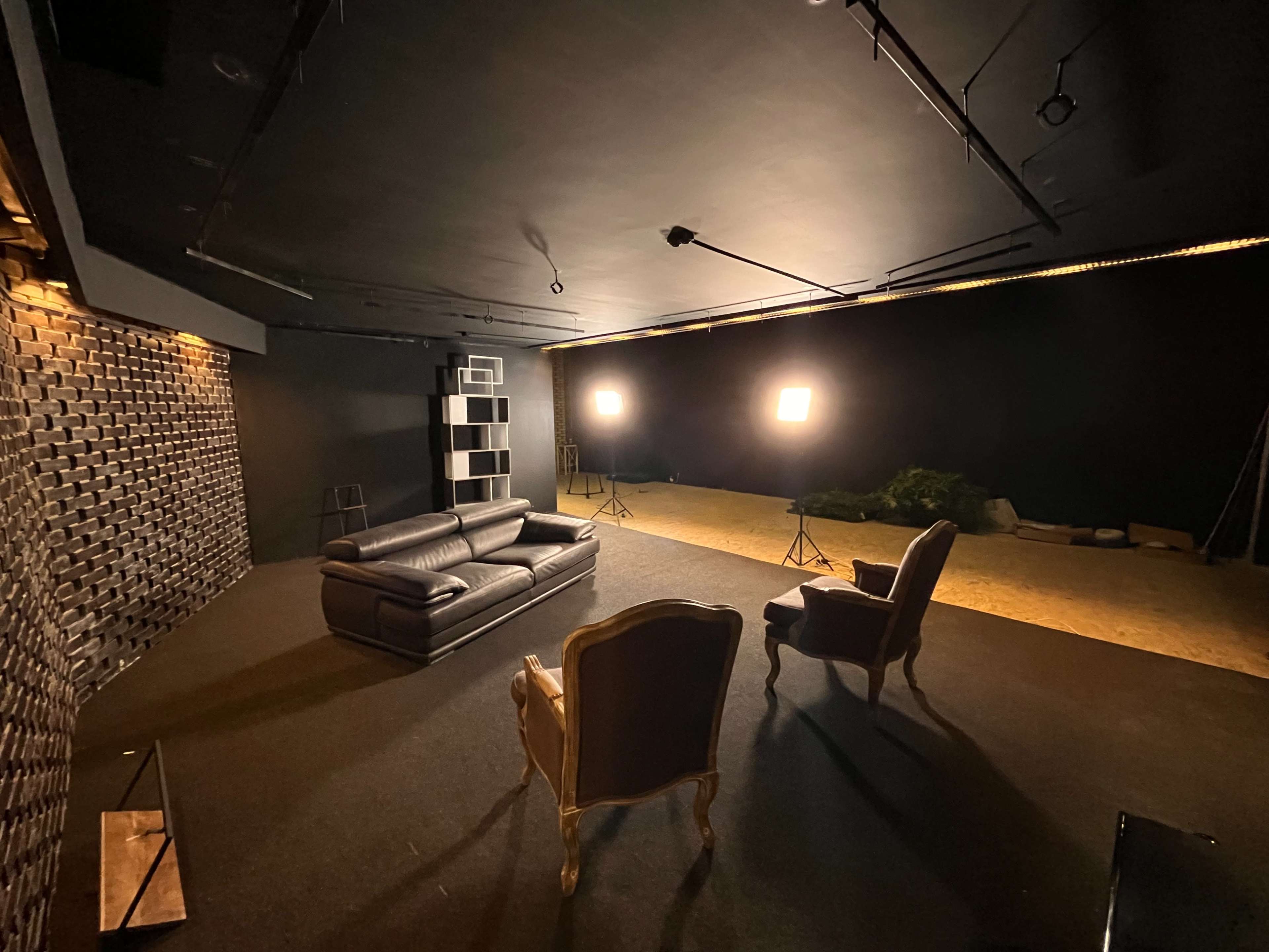 The image shows a dimly lit studio with a black wall, two spotlights illuminating a brown leather sofa, and an ornate chair placed opposite it.