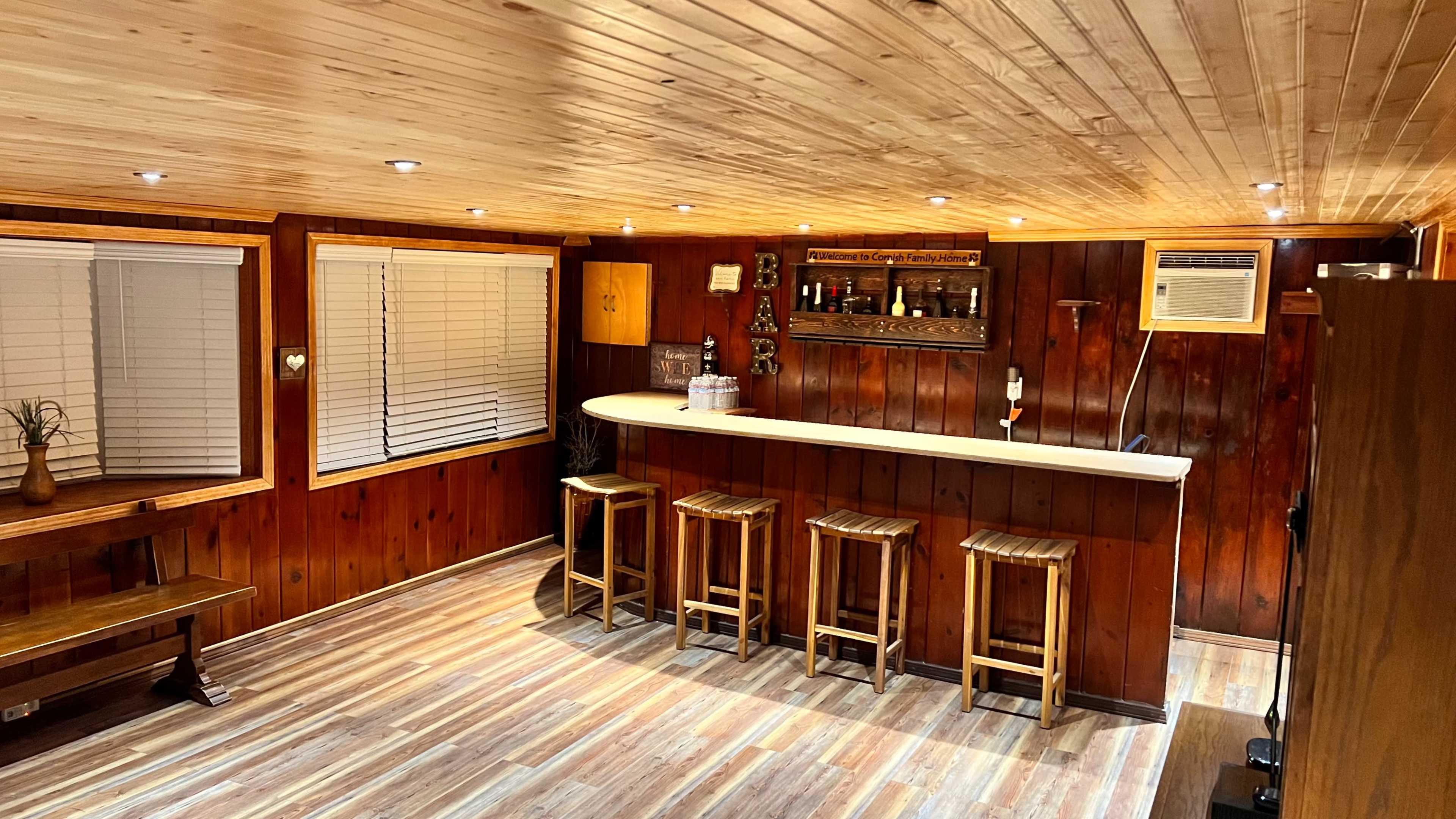 A wooden-paneled bar area with a countertop and three stools, featuring shelves with bottles in the background.