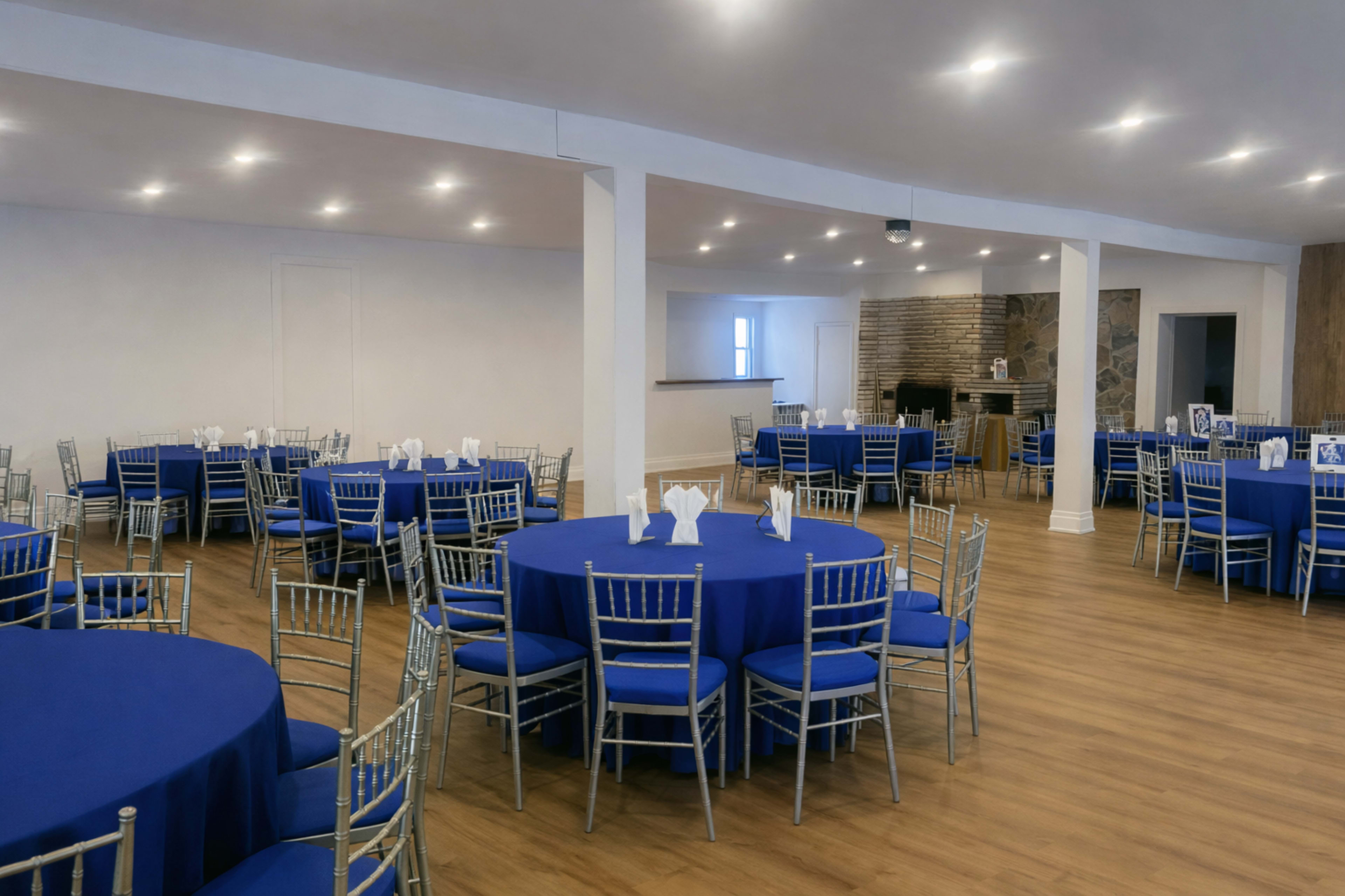 The image shows a spacious event room with several round tables covered in blue tablecloths and silver chiavari chairs arranged neatly across a wooden floor.