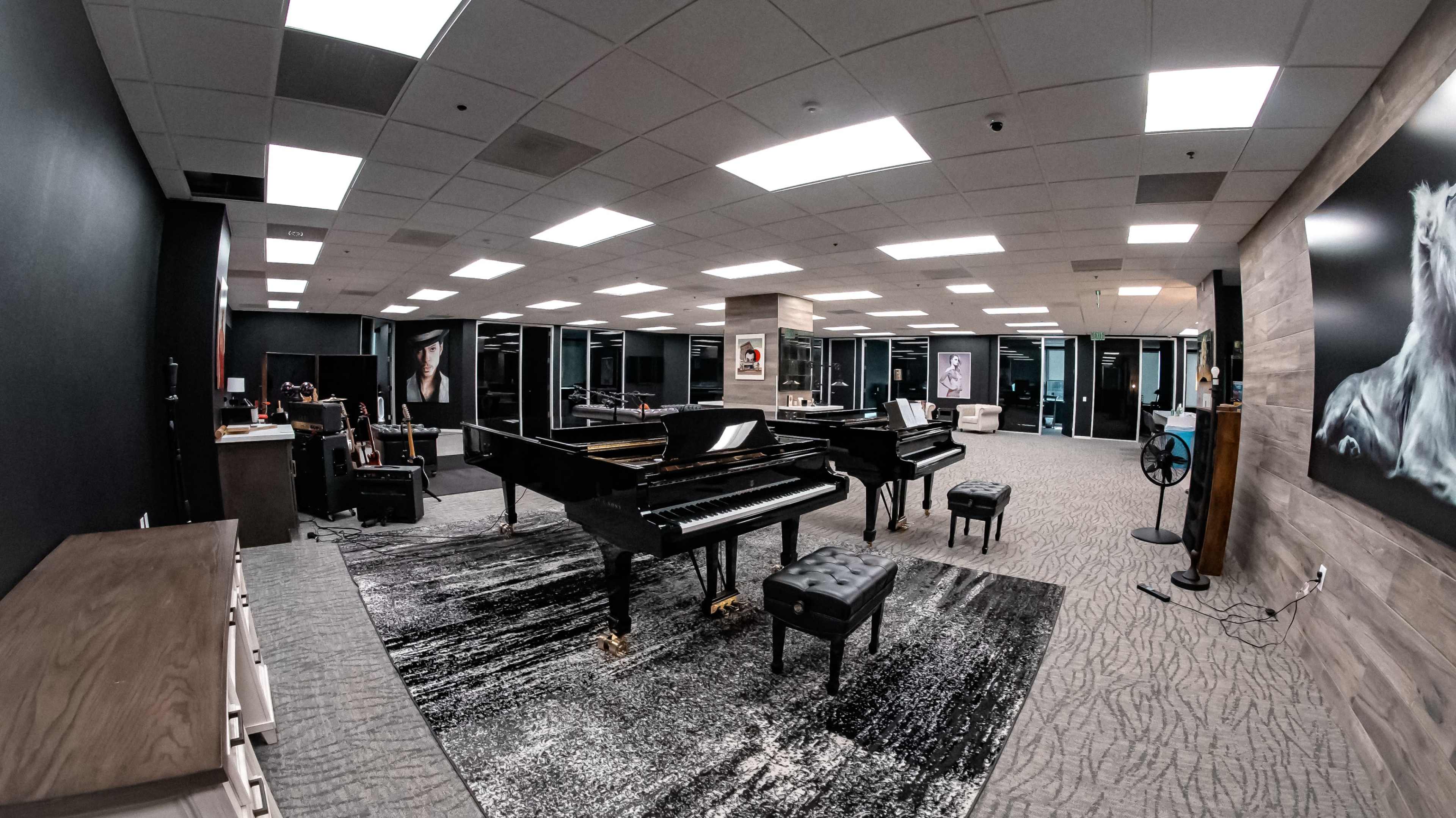 A spacious music studio featuring two grand pianos, a black rug, and large windows with various furniture arranged around the room.