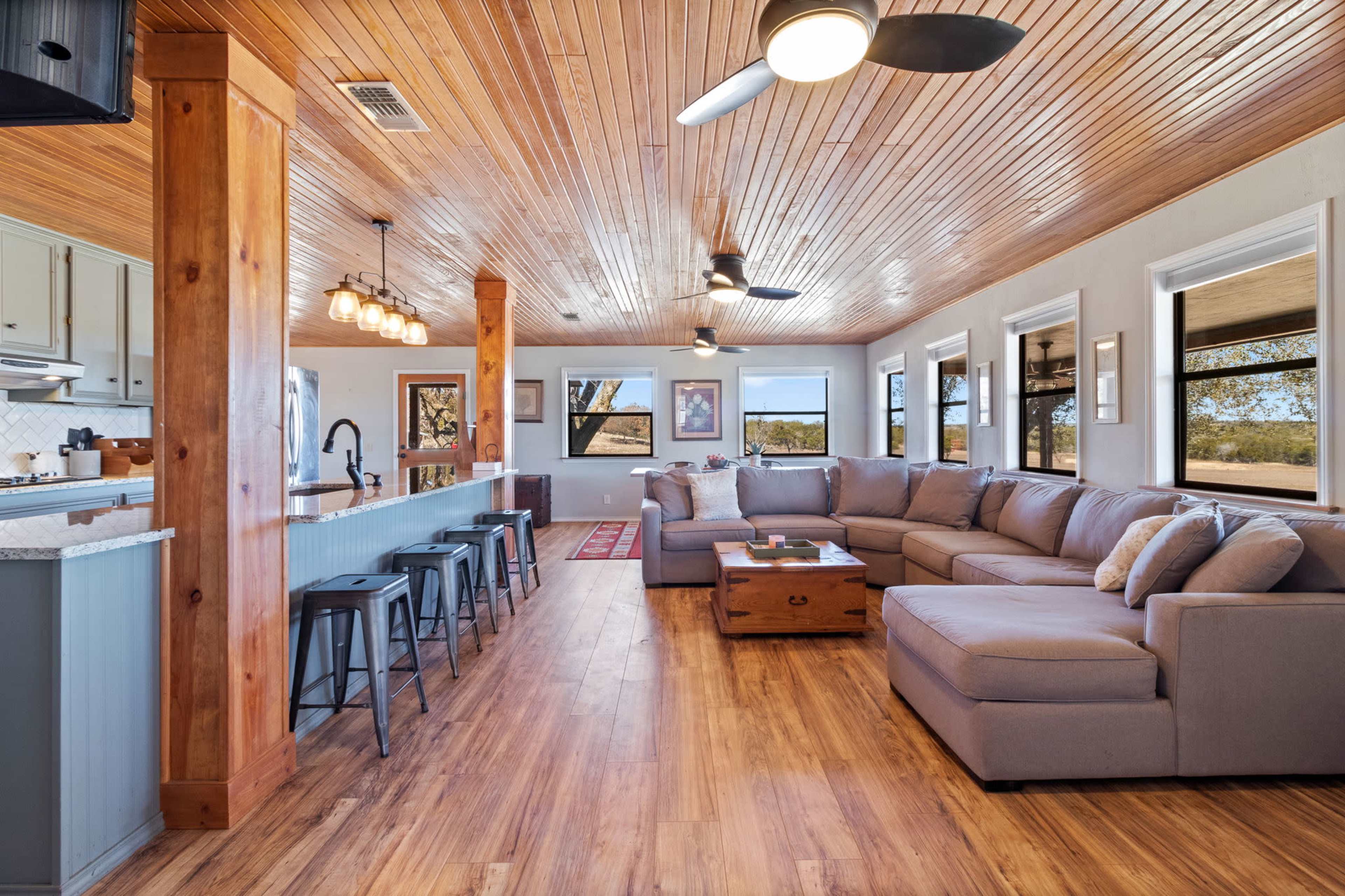 The image shows a spacious living area featuring a gray sectional sofa, a wooden coffee table, and large windows that provide ample natural light.