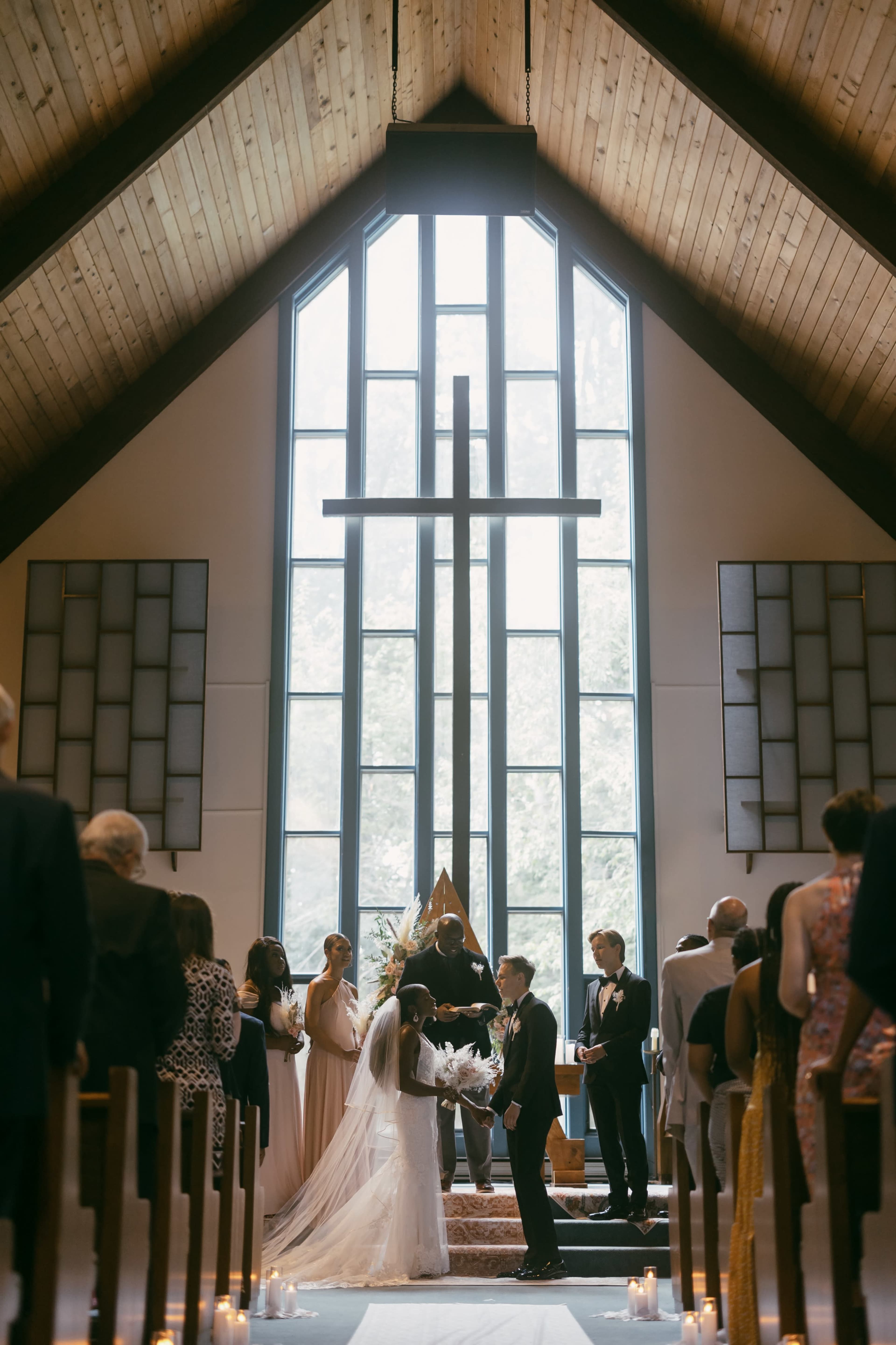 A bride and groom stand under a large cross during their wedding ceremony in a church, surrounded by guests and bridal party members.