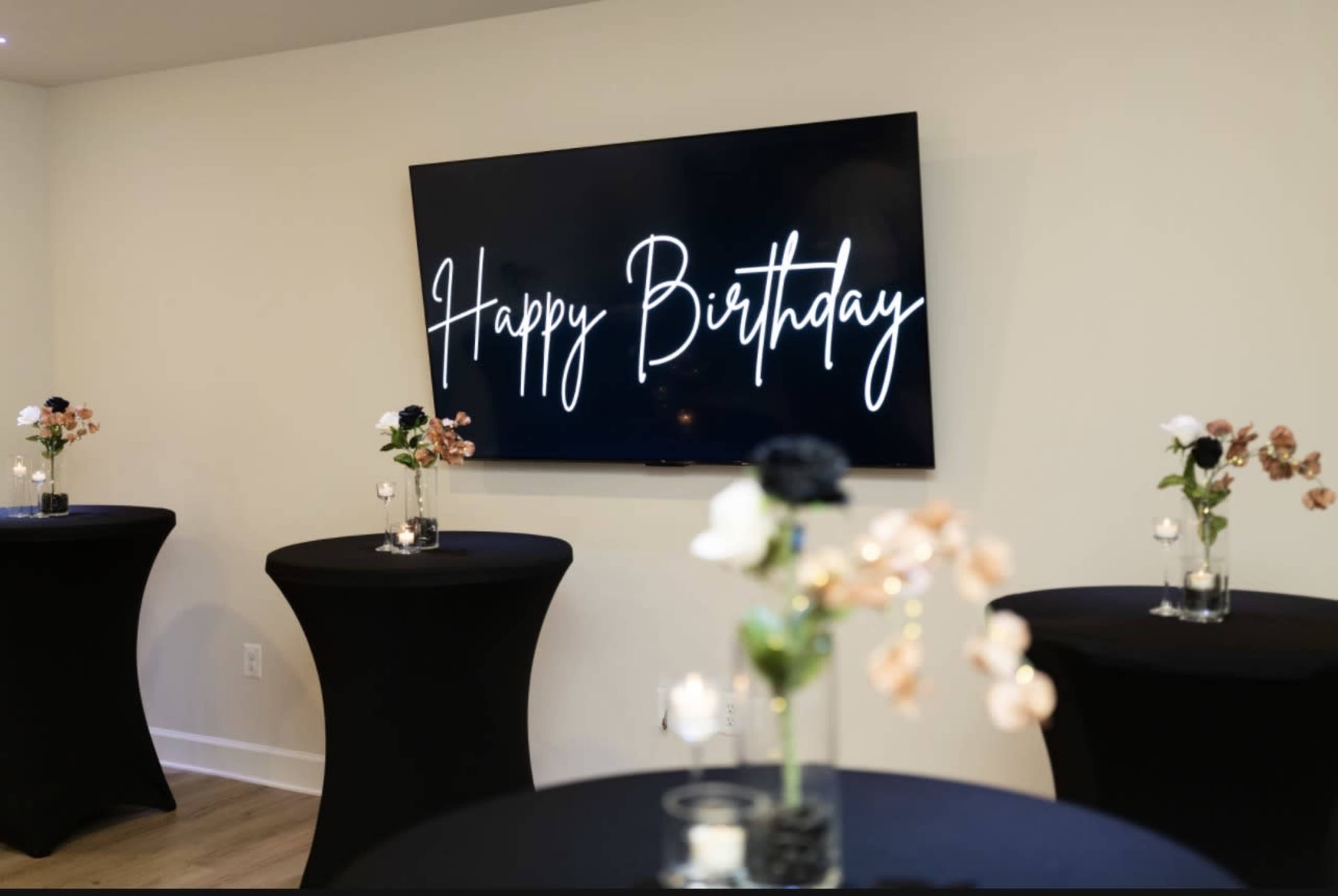 The image shows a room adorned with black tables topped with floral arrangements, featuring a large screen displaying the text "Happy Birthday."