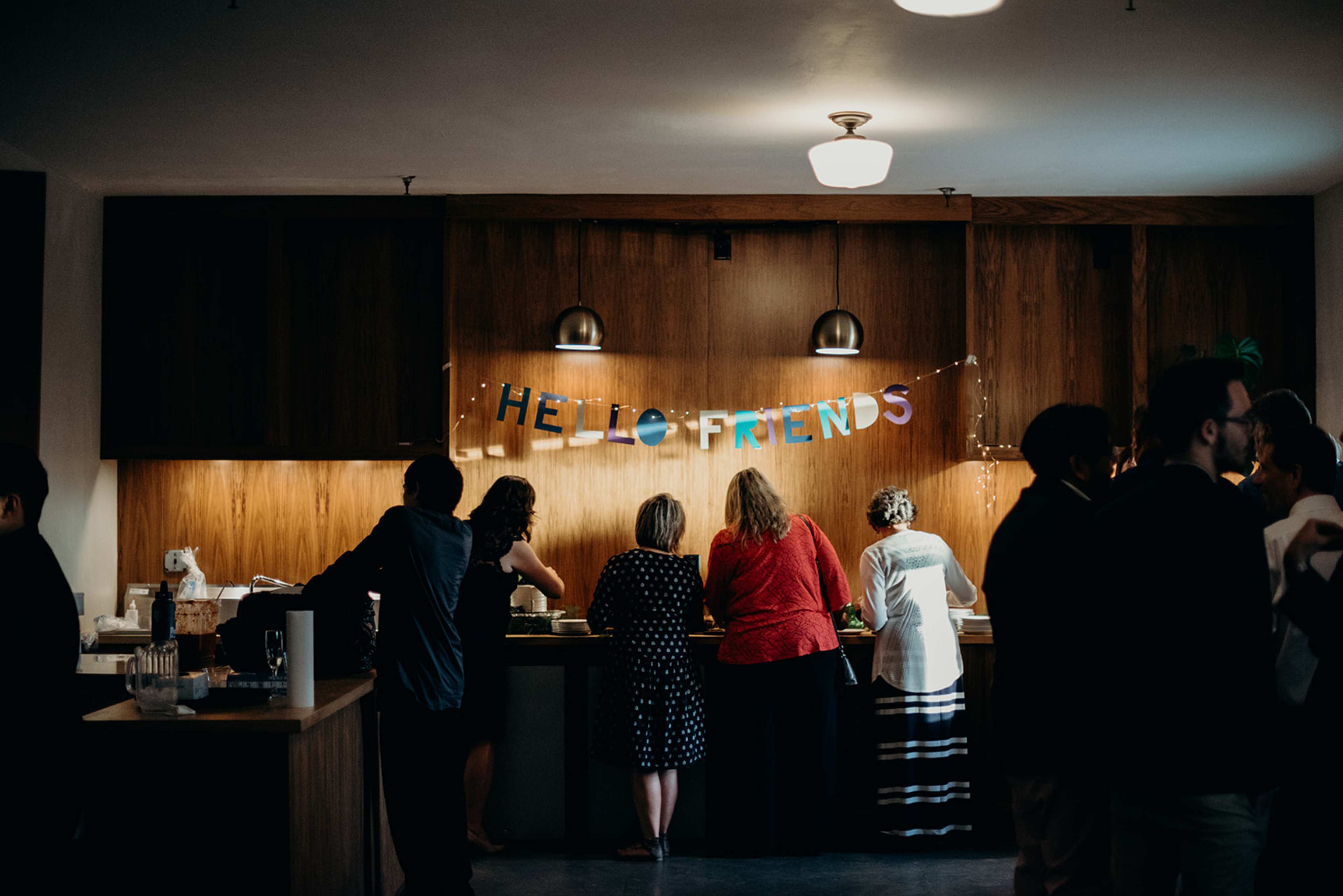 A group of people stands near a bar area with a "HELLO FRIENDS" banner displayed on the wall.