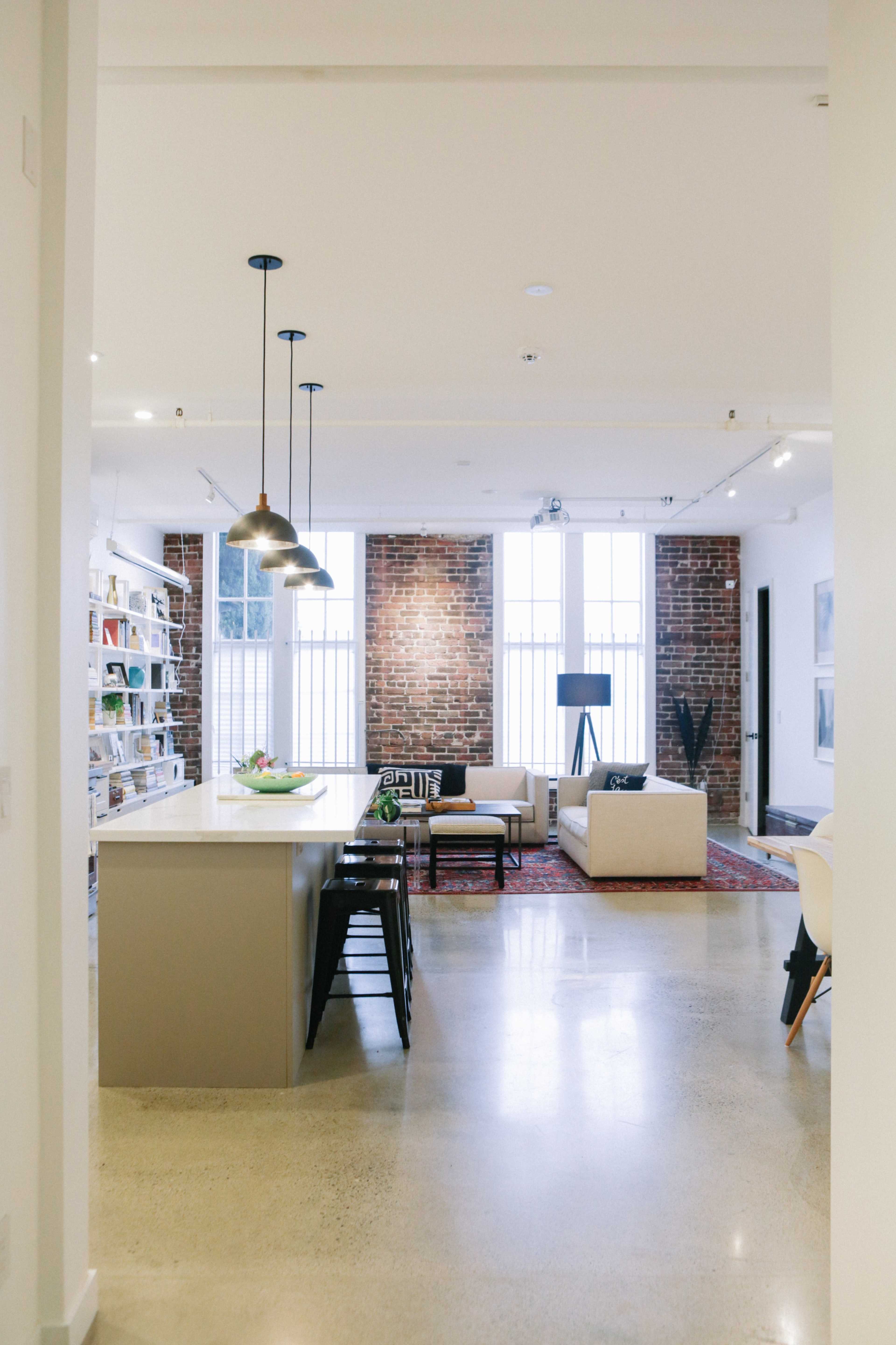 A modern open-concept living space featuring a kitchen with a countertop, black chairs, and a cozy seating area with a sofa and decorative rug against a brick wall.
