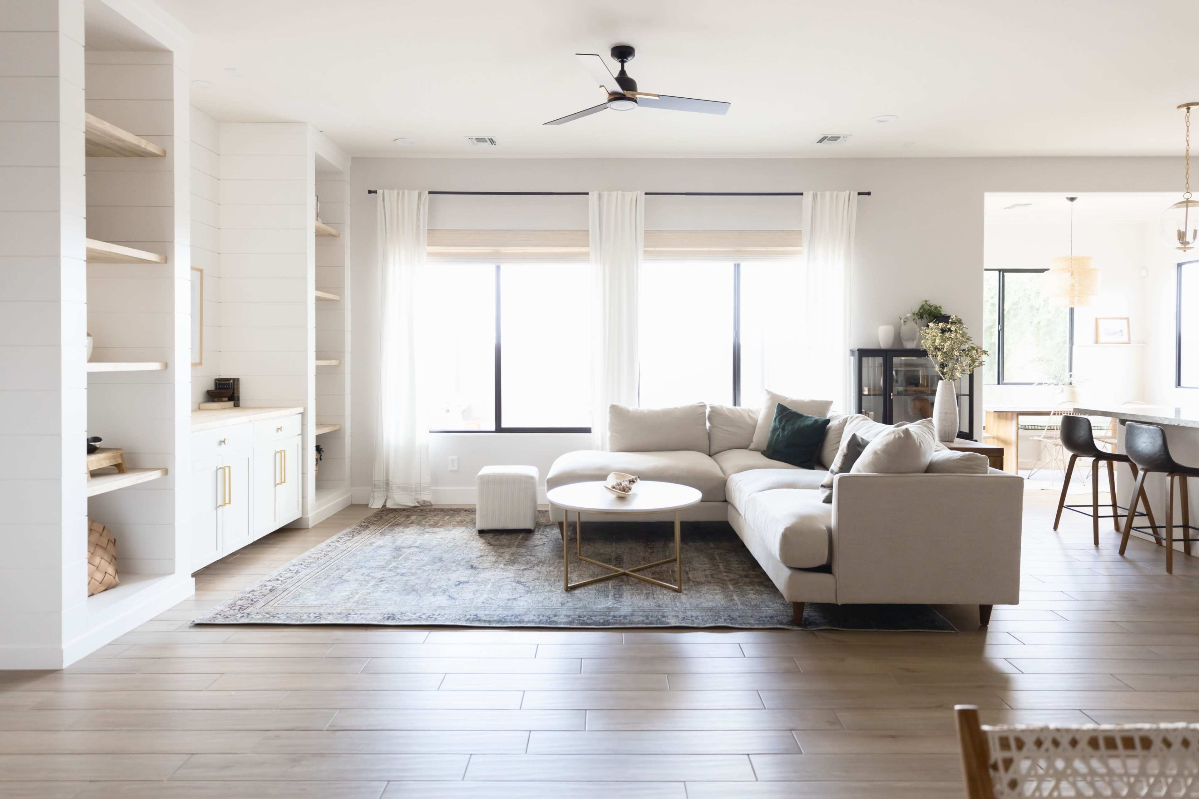 A bright, modern living room with a sectional sofa, an area rug, large windows, and a dining area visible in the background.