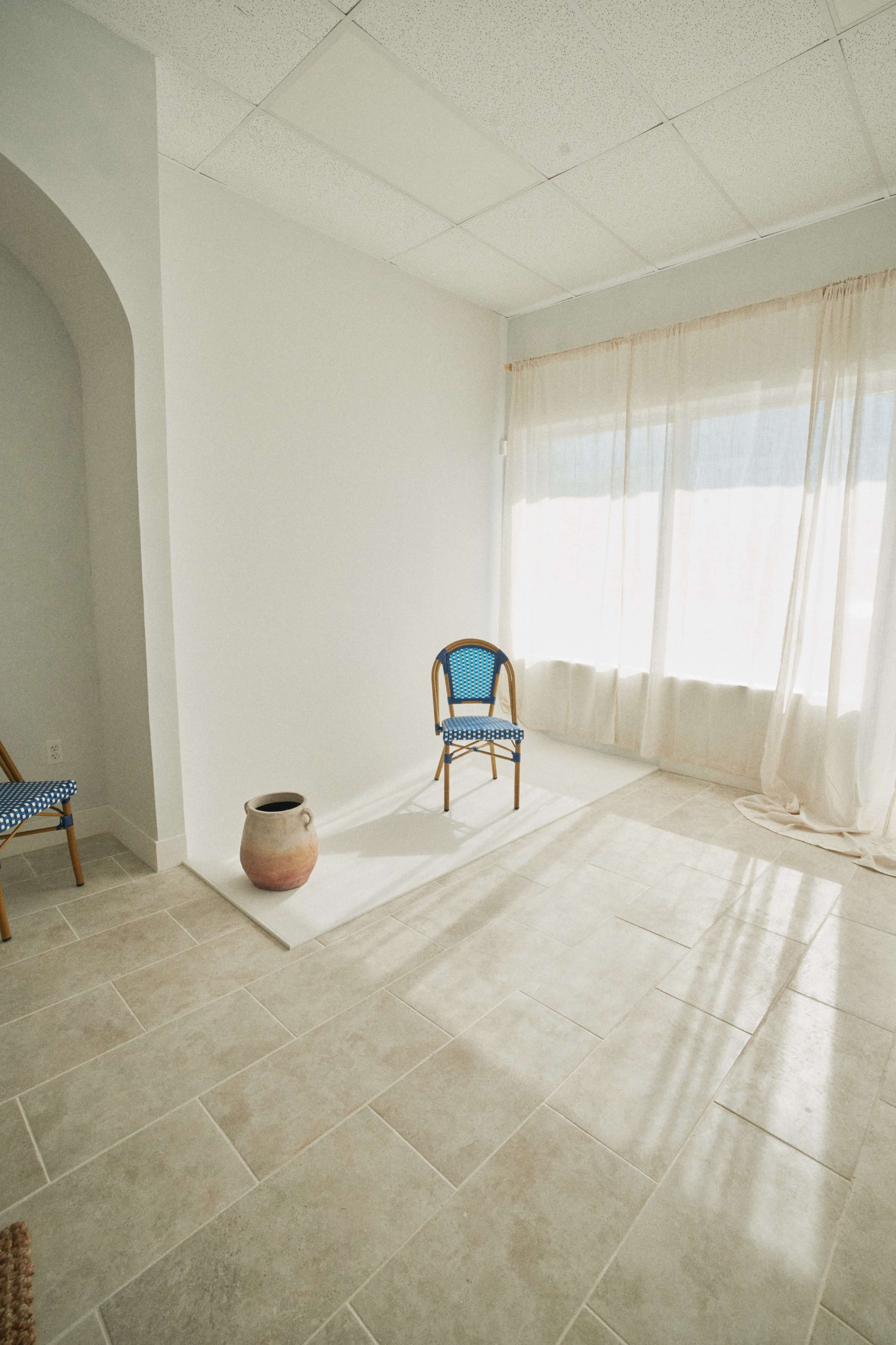 The image shows a minimalist room with a single chair and a pottery vase, illuminated by natural light from a large window.
