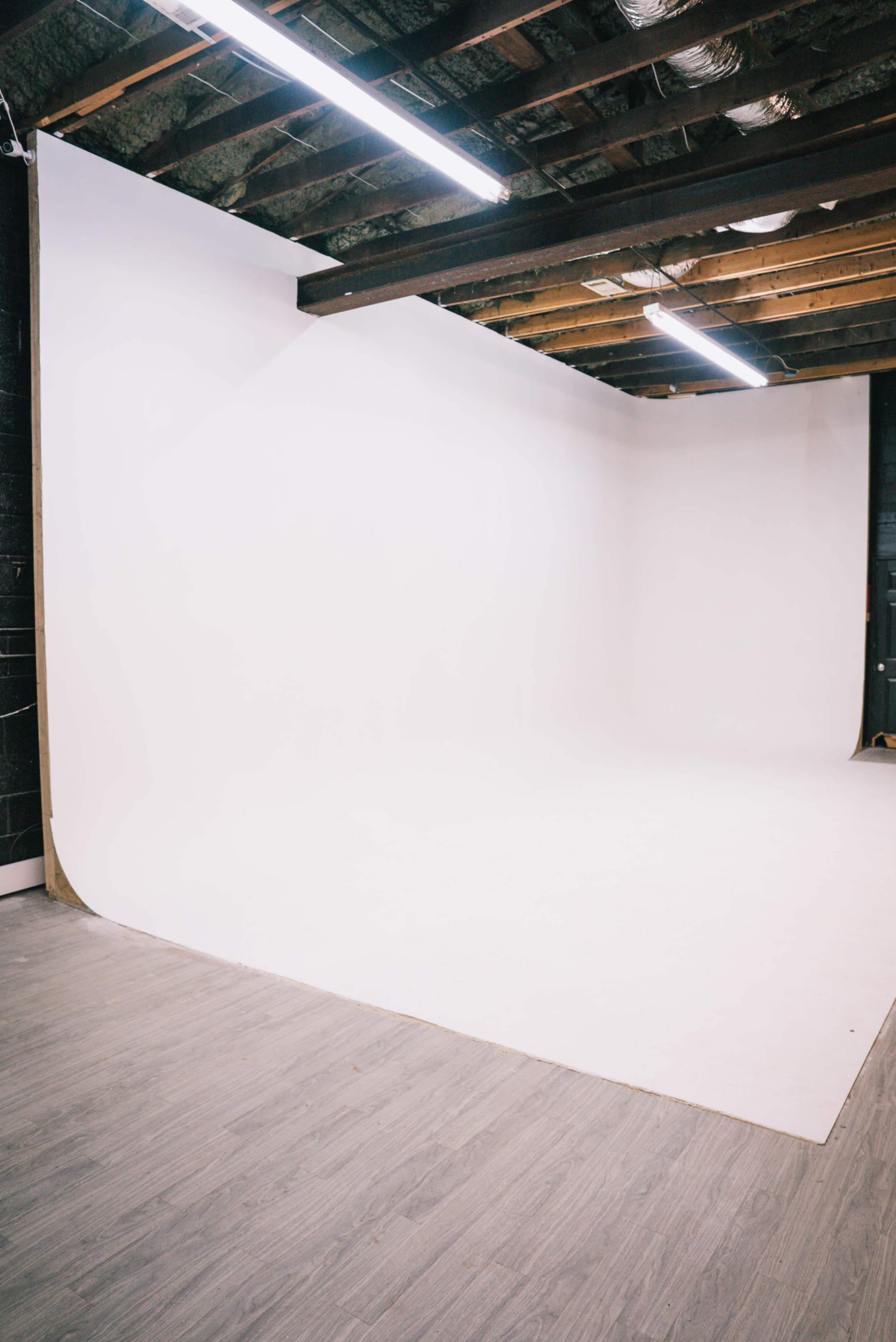 The image shows a minimalist photography studio with a large white backdrop and wooden beams on the ceiling.