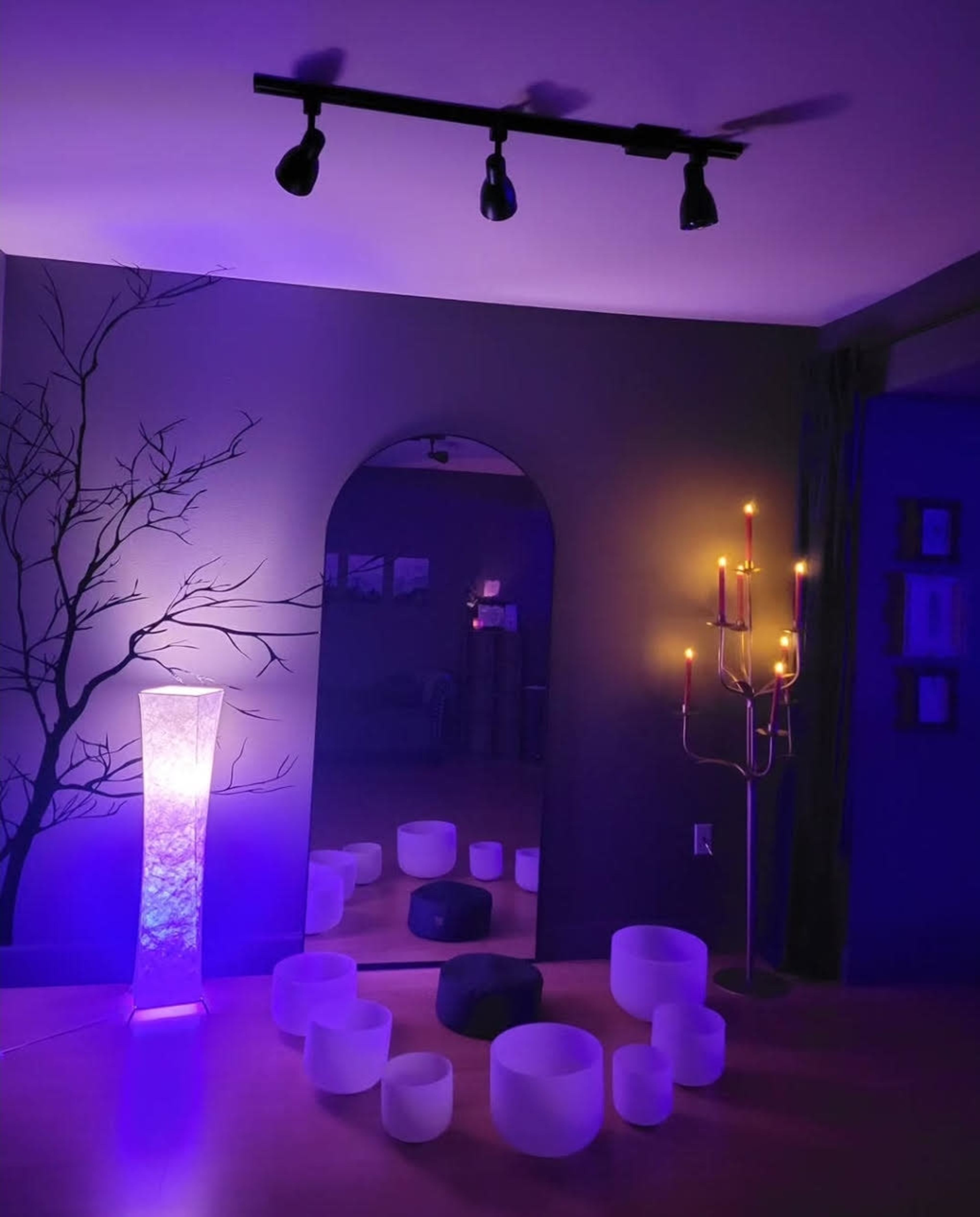 The scene features a dimly lit room with purple lighting, an arch-shaped mirror, a floor lamp, several crystal bowls arranged on the floor, and a candle holder with lit candles.