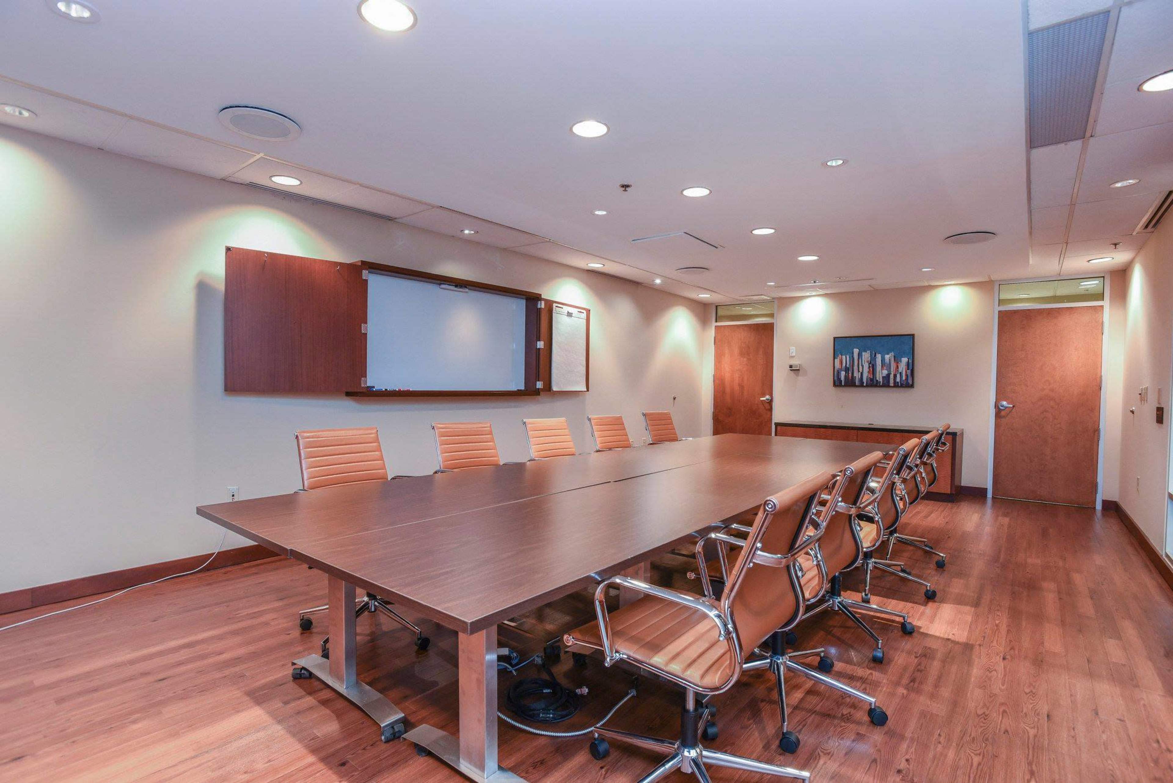 A large conference room features a long wooden table surrounded by several orange chairs, with a whiteboard and wall-mounted artwork.