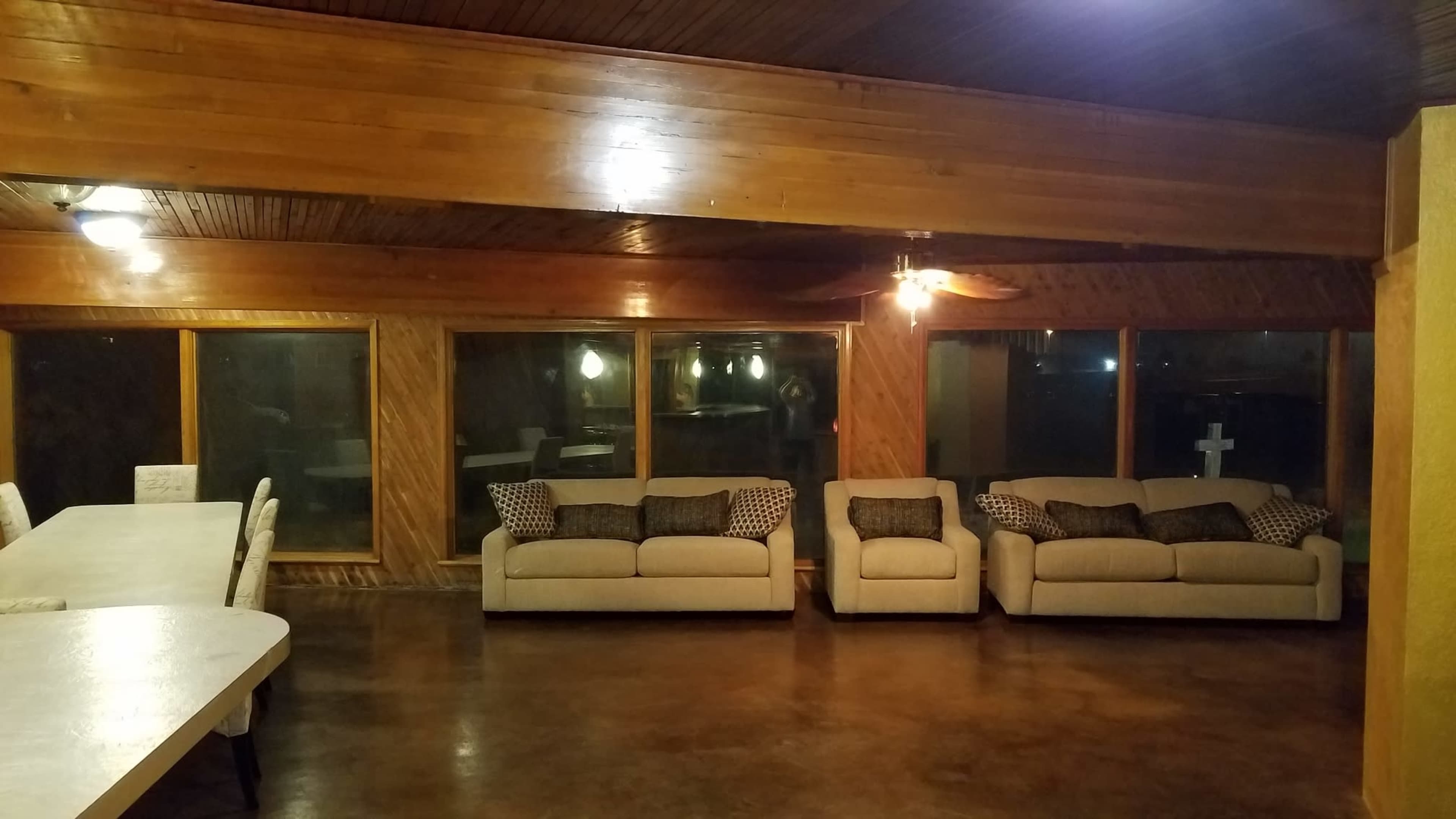 A spacious interior with wooden walls and a polished concrete floor, featuring two beige sofas with cushions and large windows that overlook a dark outdoor area.