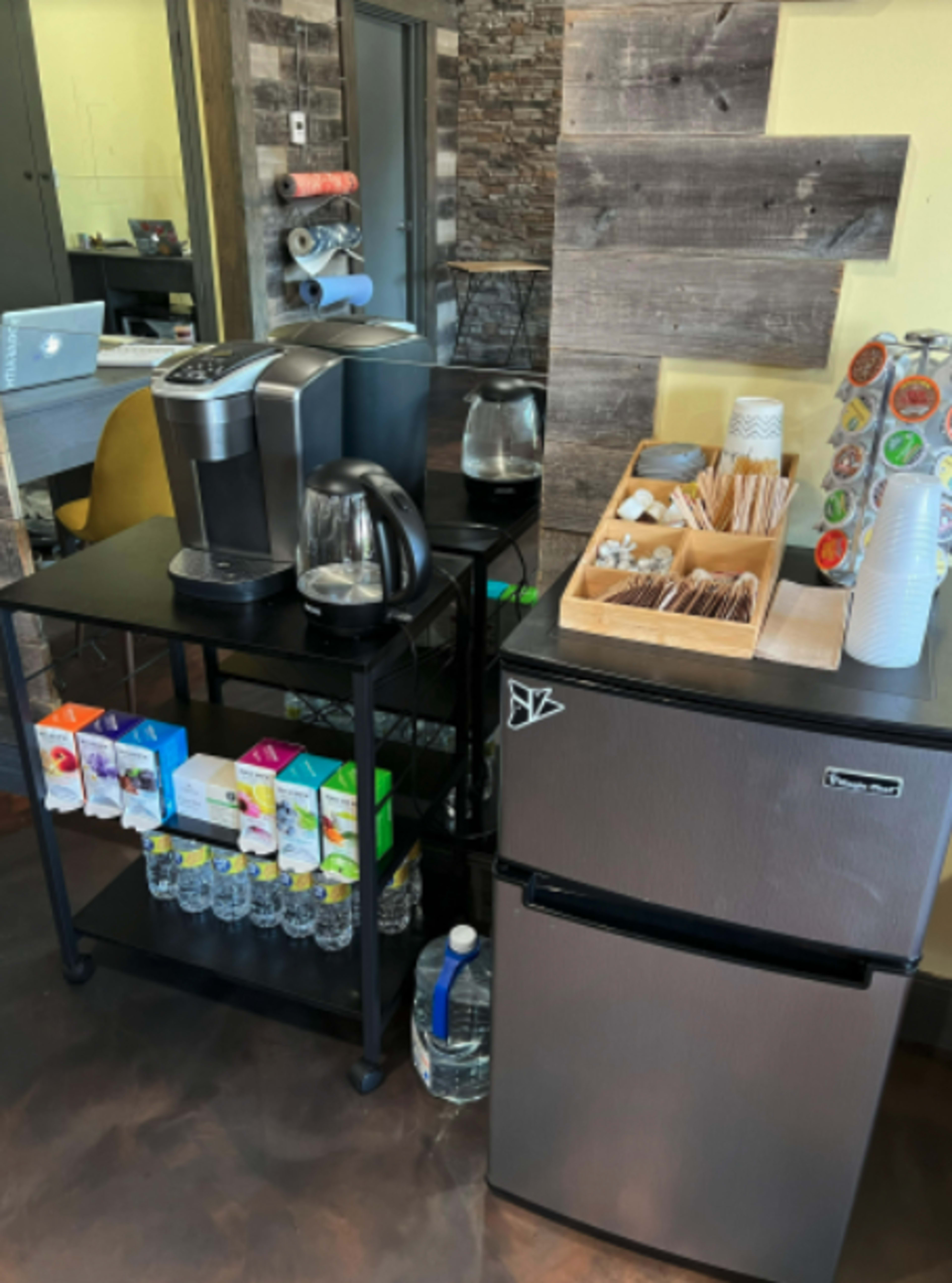 A coffee station with a coffee maker, water bottles, and a mini fridge next to a wooden wall.