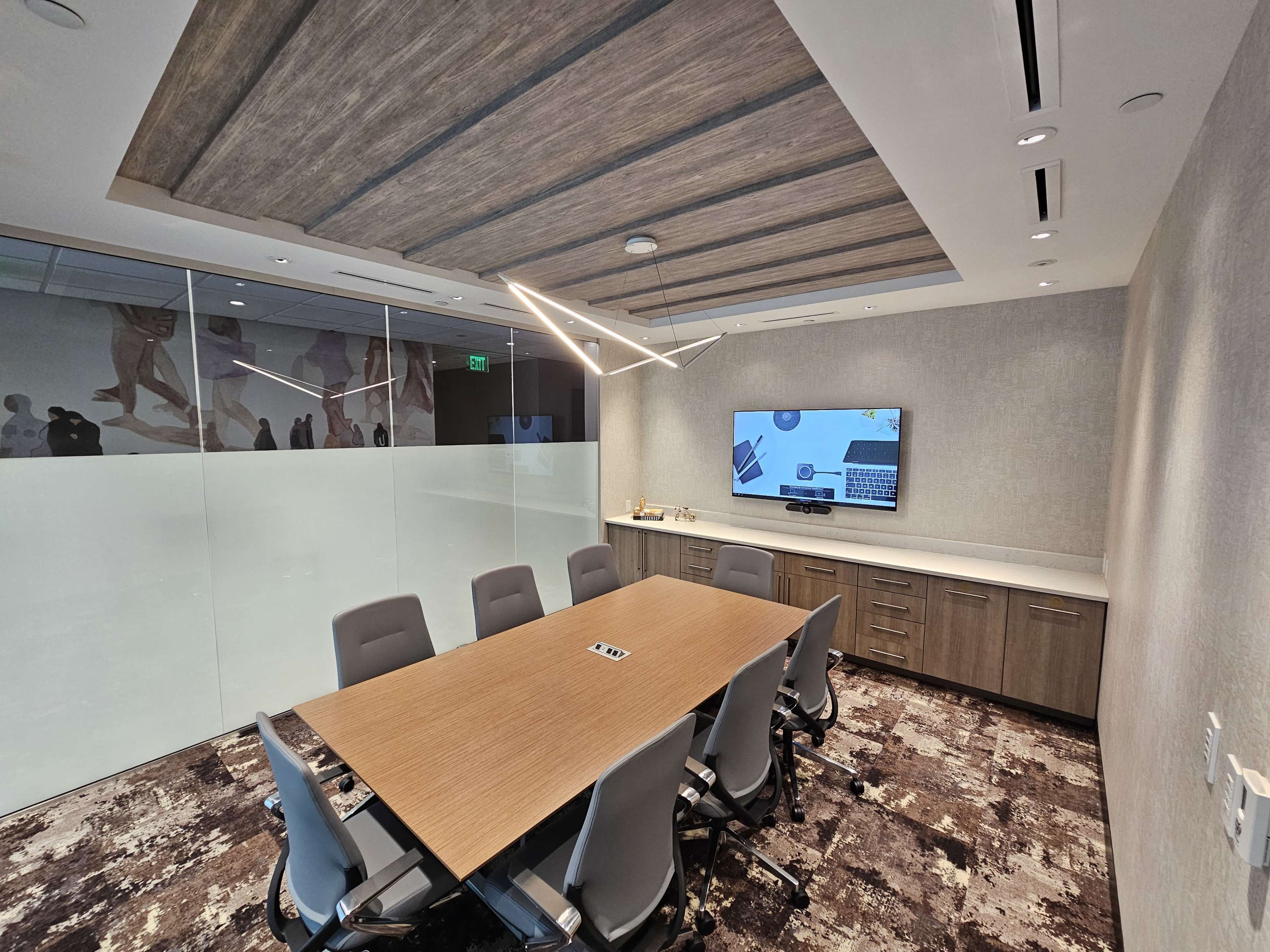 A modern conference room with a large wooden table, ergonomic chairs, and a wall-mounted screen displaying a computer interface.