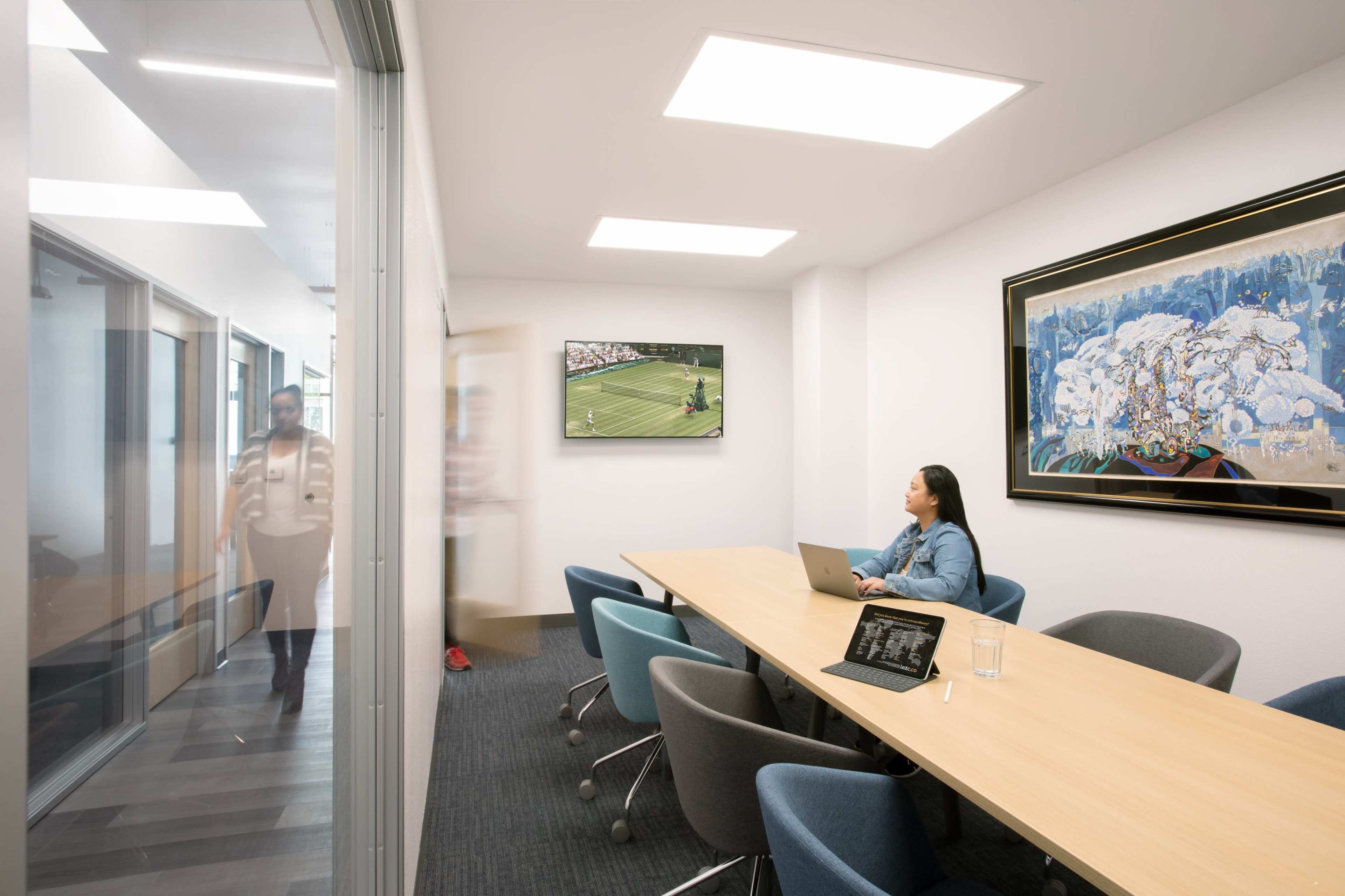 A woman works on a laptop at a long wooden table in a brightly lit conference room, while a person walks through an adjacent glass door.