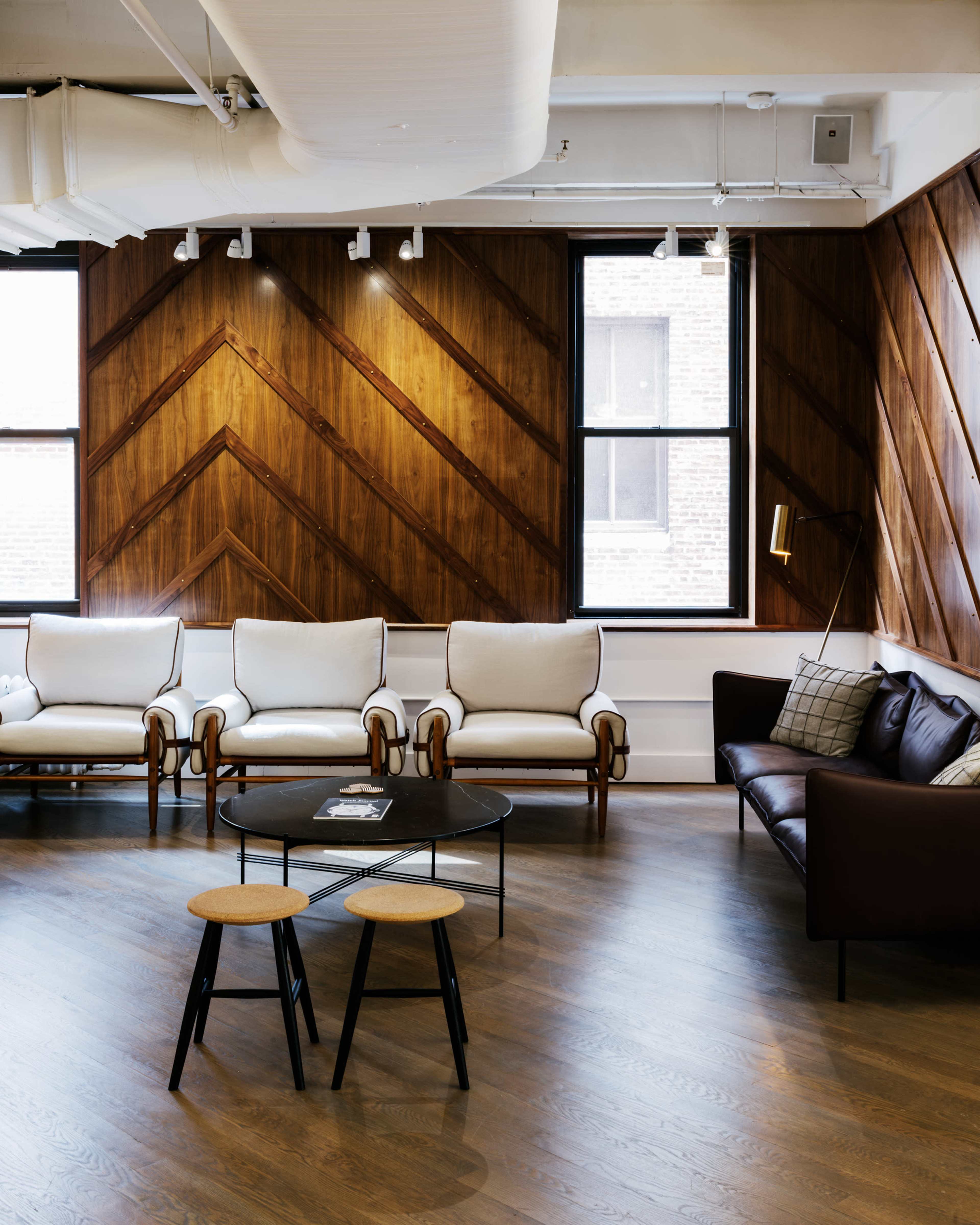 The image shows a modern lounge area featuring a set of white armchairs, a dark sofa, and a round coffee table between two small stools, all set against a wooden paneled wall.