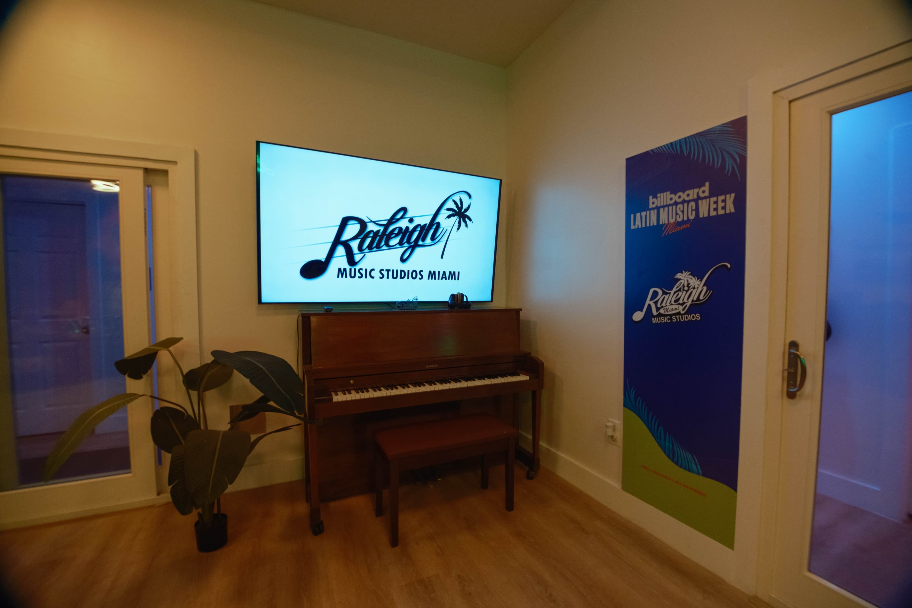 The image shows a modern music studio with a piano, a large screen displaying "Raleigh Music Studios Miami," and a wall featuring a promotional banner for "Billboard Latin Music Week."