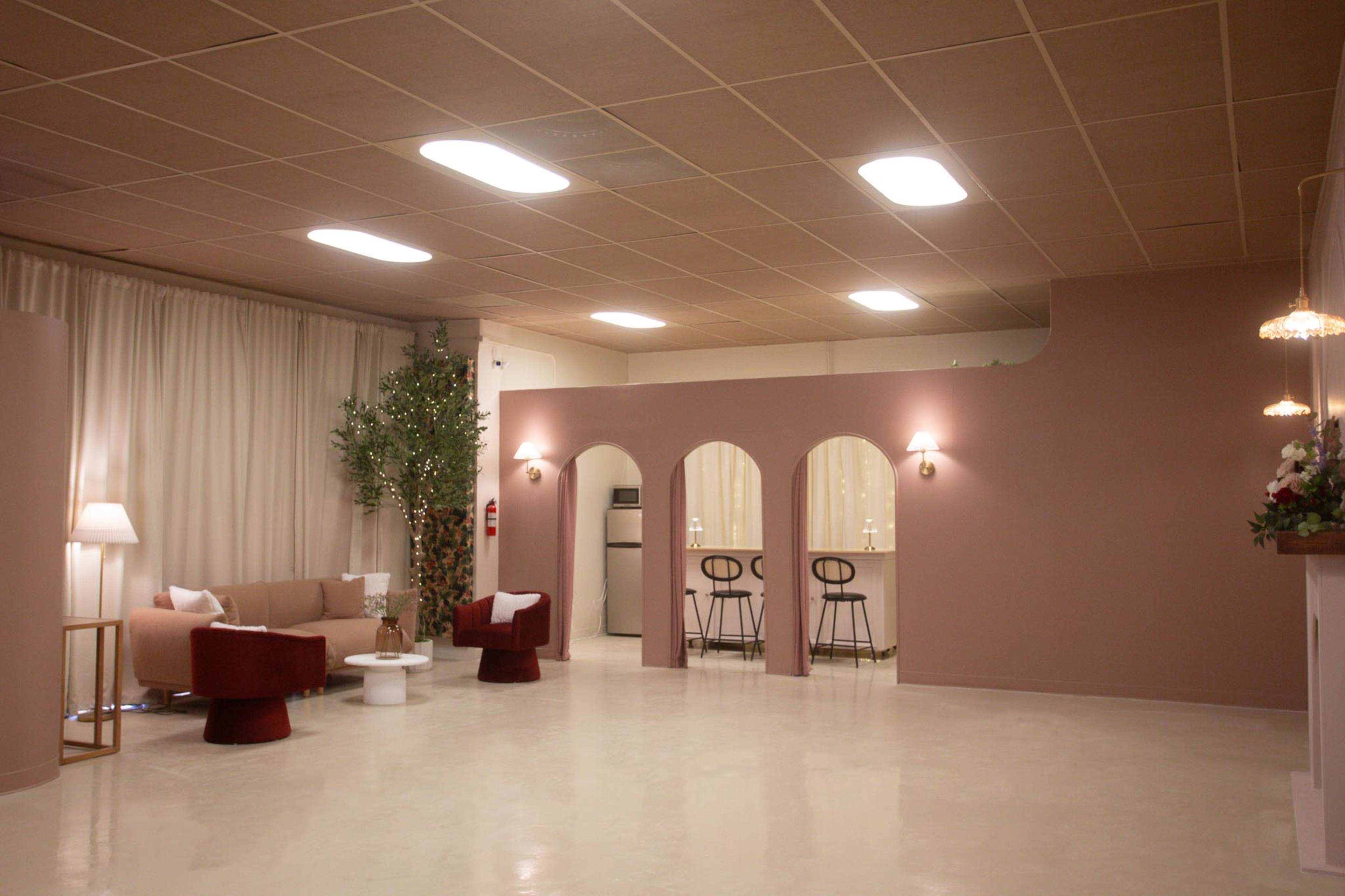 A modern indoor space with a seating area, featuring a beige sofa, red chairs, and a small table, alongside a bar area with stools against a pink wall.