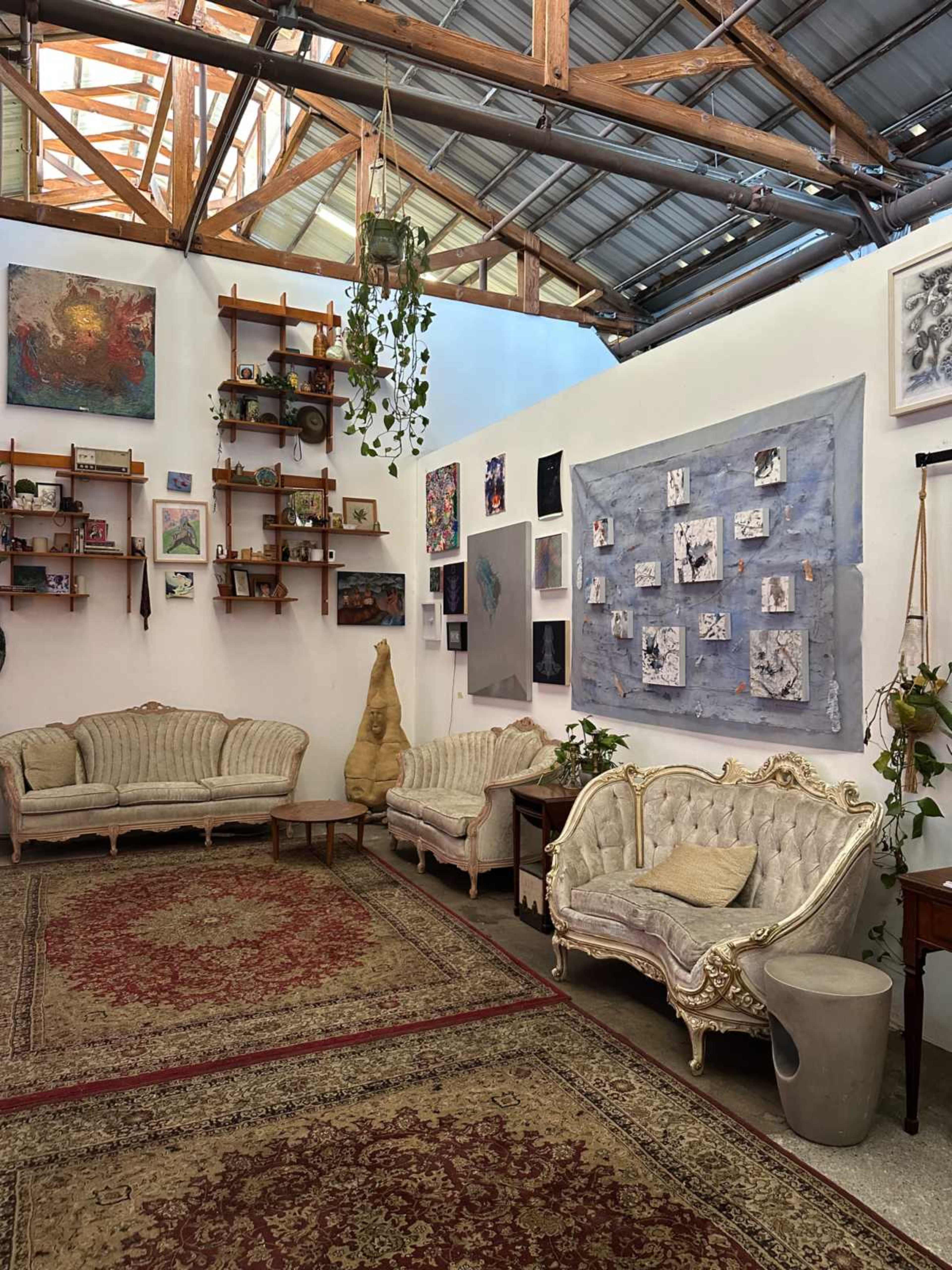 The image shows a spacious room with vintage-style sofas, a patterned rug, and an assortment of artwork displayed on the walls along with shelves filled with various items.