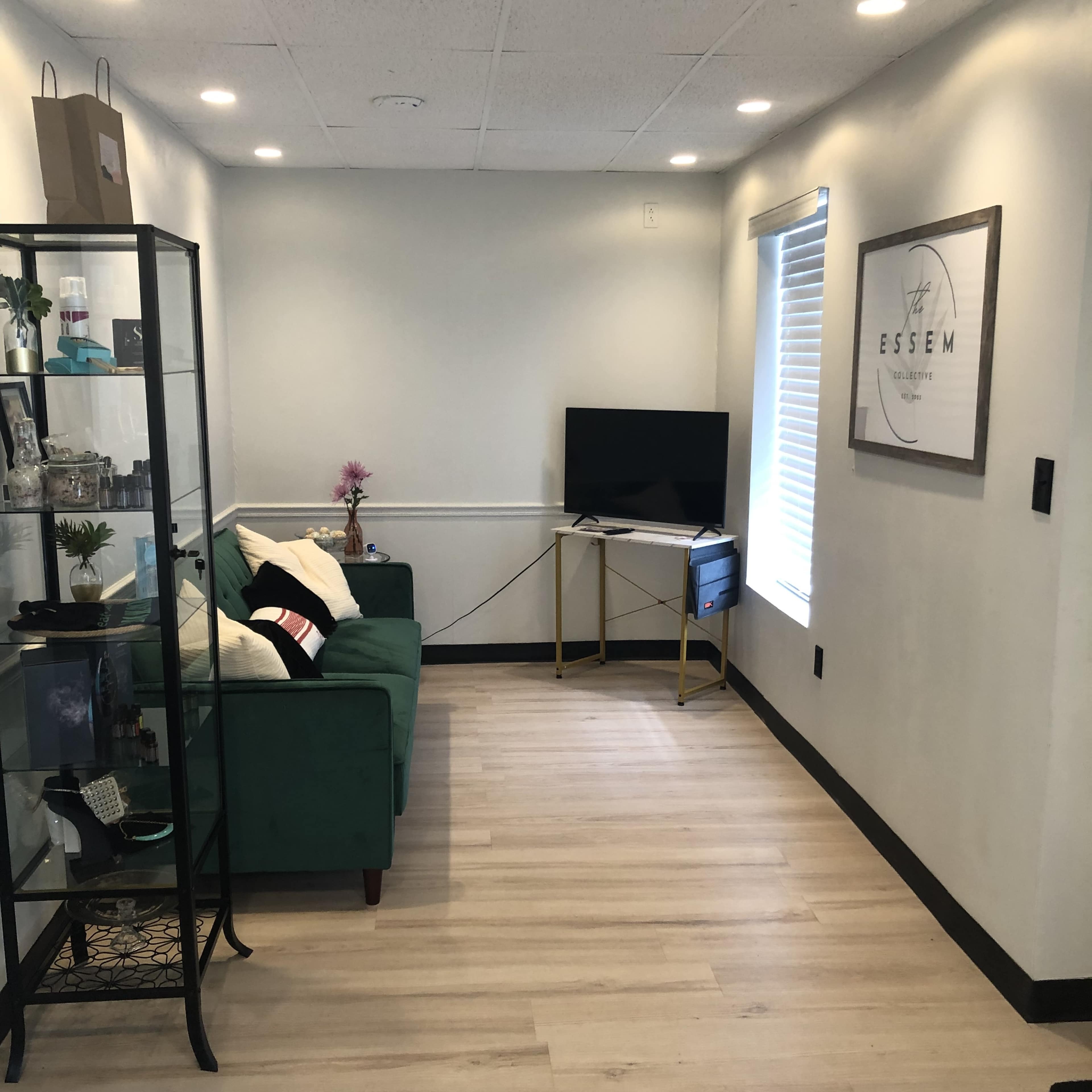 The image shows a small room with a green sofa, a black shelving unit displaying various items, a television on a stand, and a window with blinds.