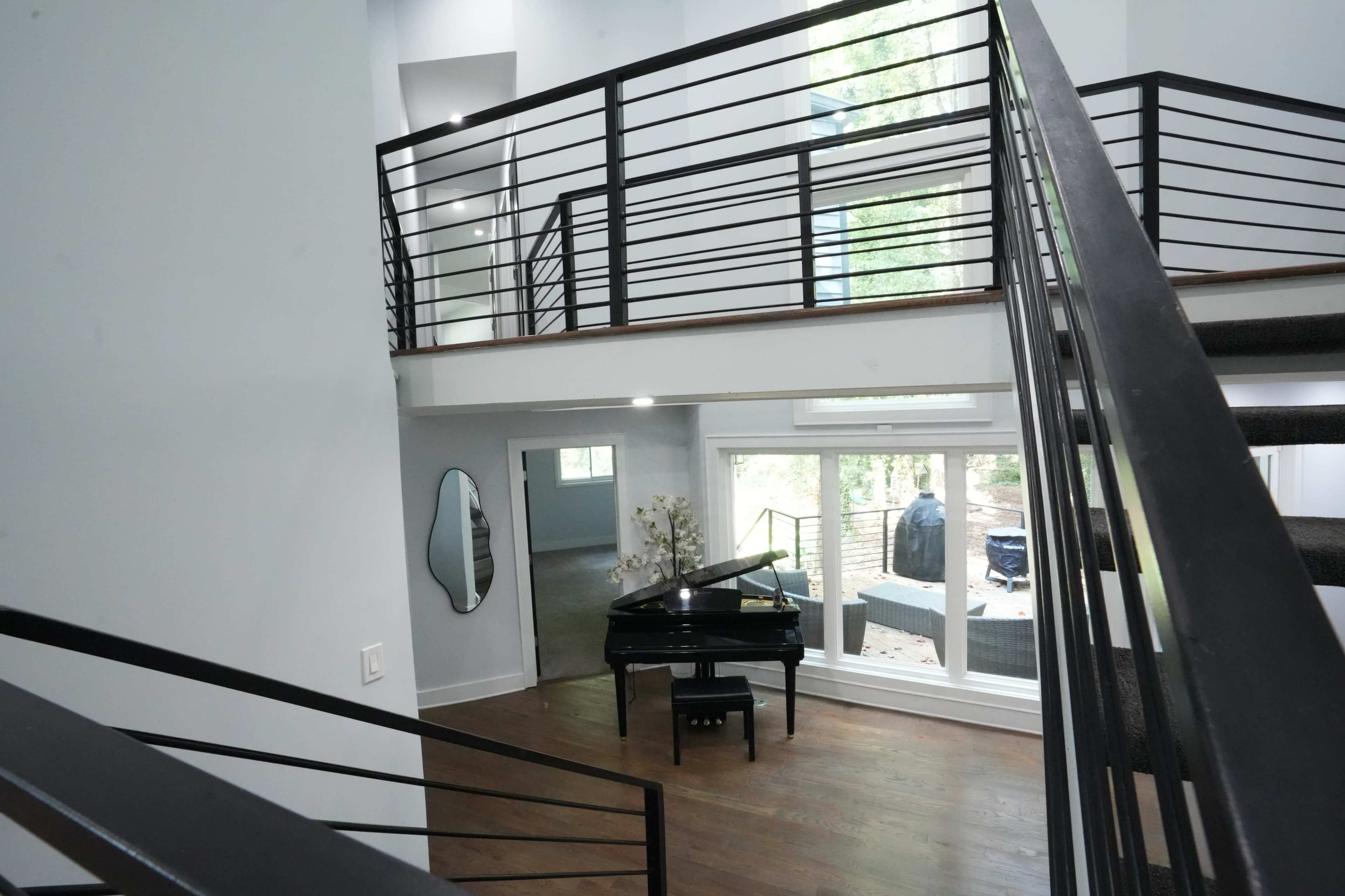 The image shows a modern interior space featuring an open balcony area overlooking a living room with a black piano and large windows allowing natural light.