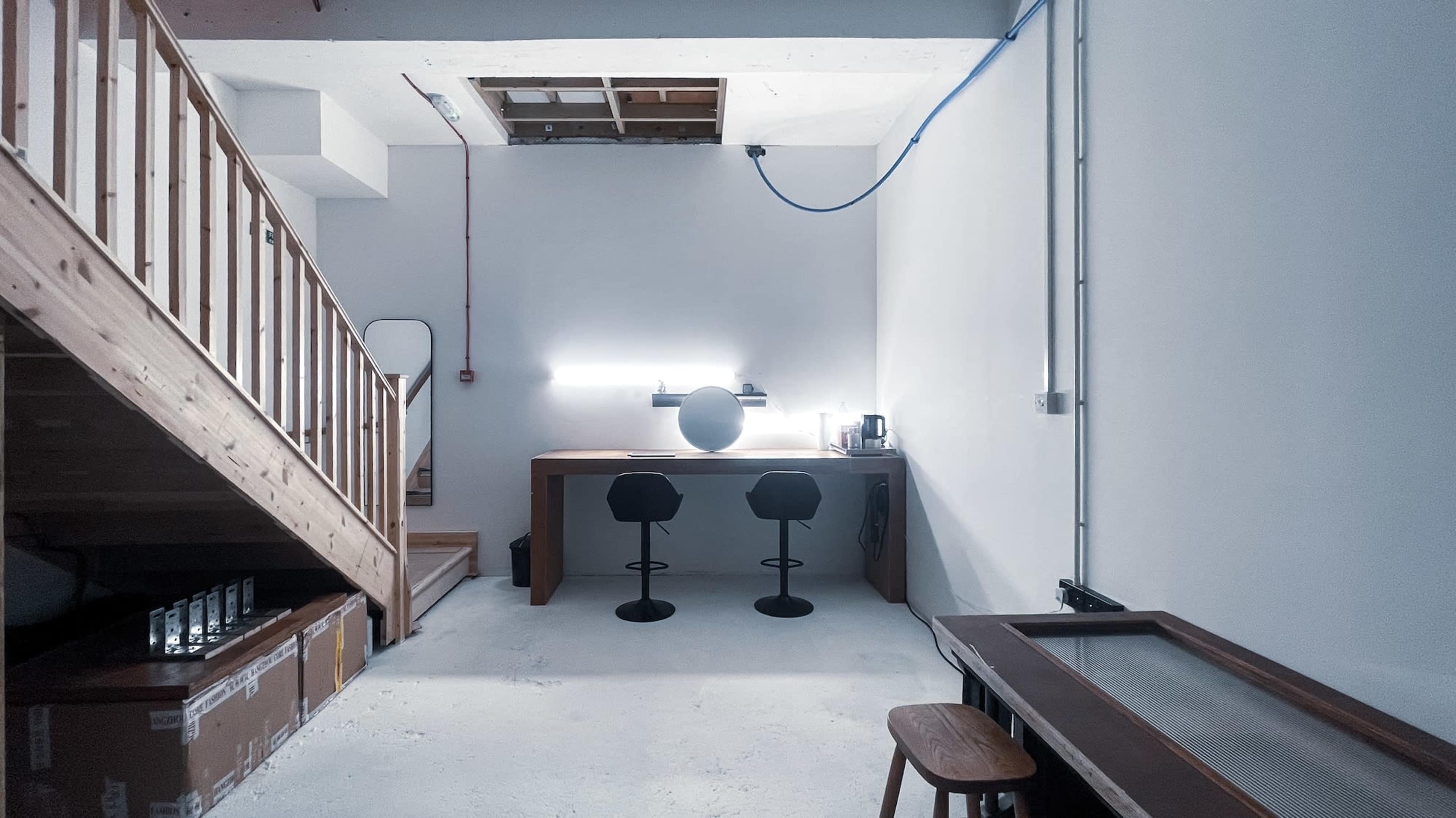The image shows a minimalistic workspace in a basement with a wooden staircase, a large desk, two black stools, and a round lamp.
