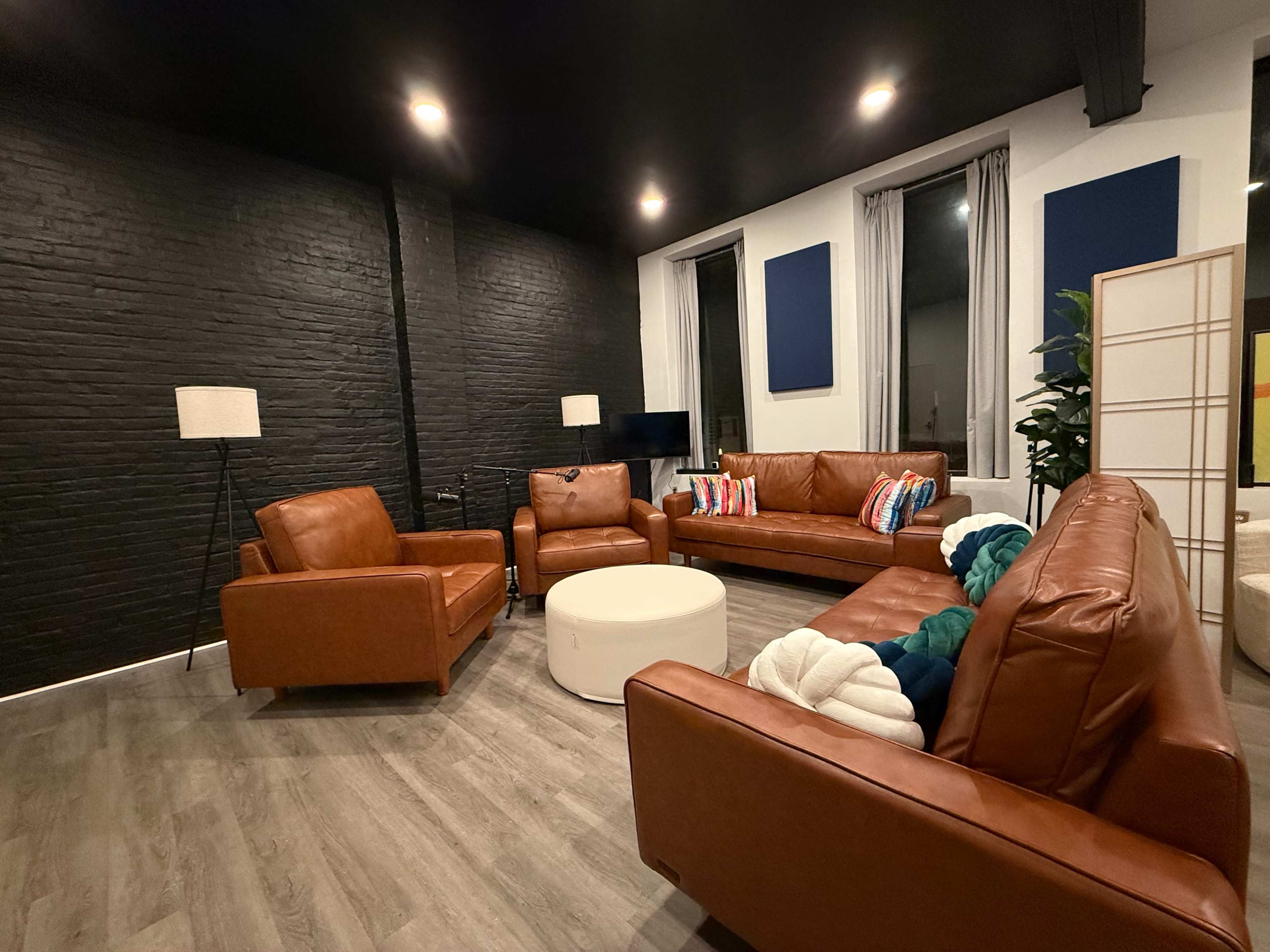 The image shows a stylish seating area featuring brown leather sofas and armchairs surrounding a white coffee table, with black walls and large windows.