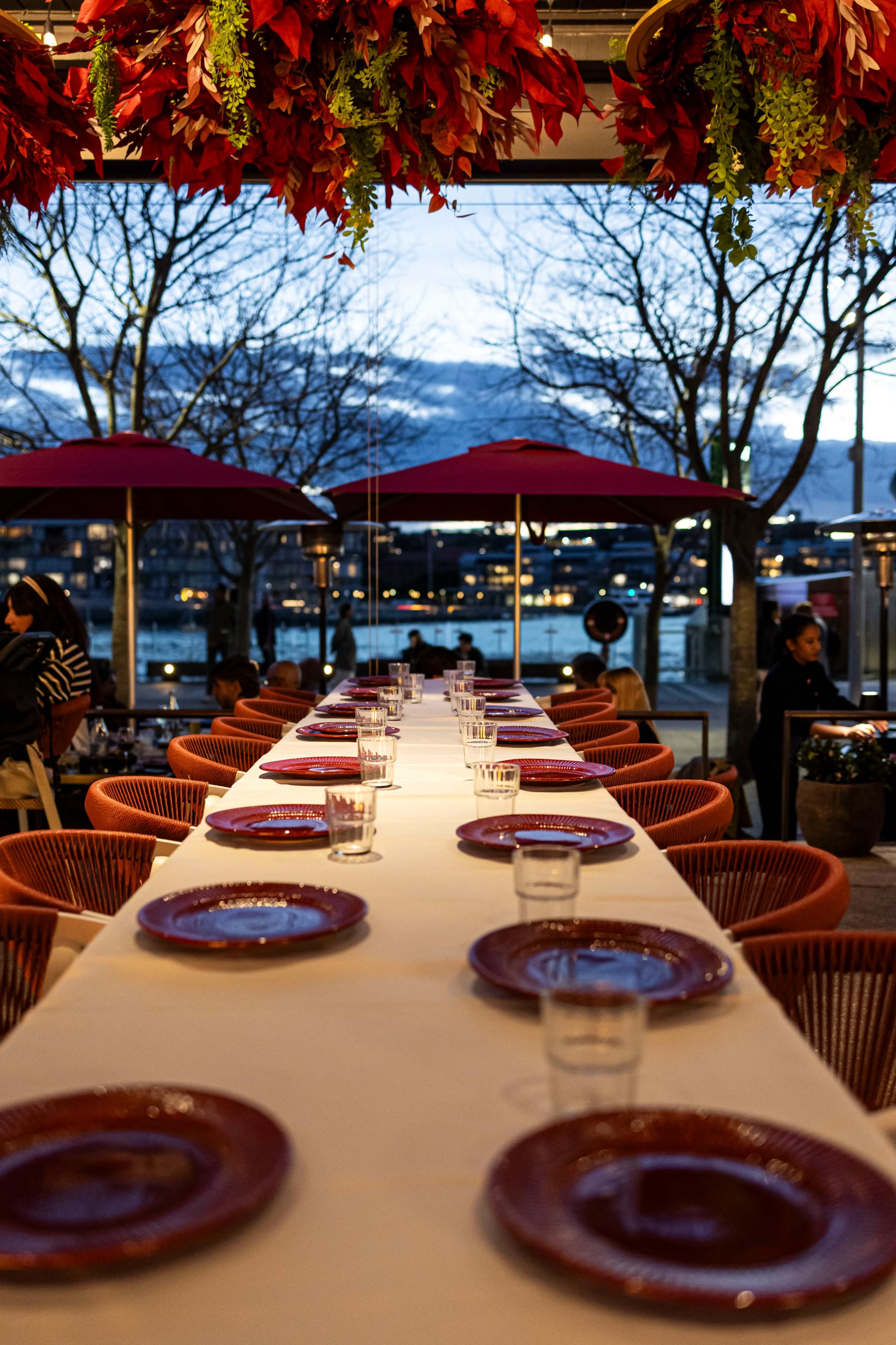 A long dining table is set with plates and glasses under red floral decorations, overlooking a waterfront view at dusk.