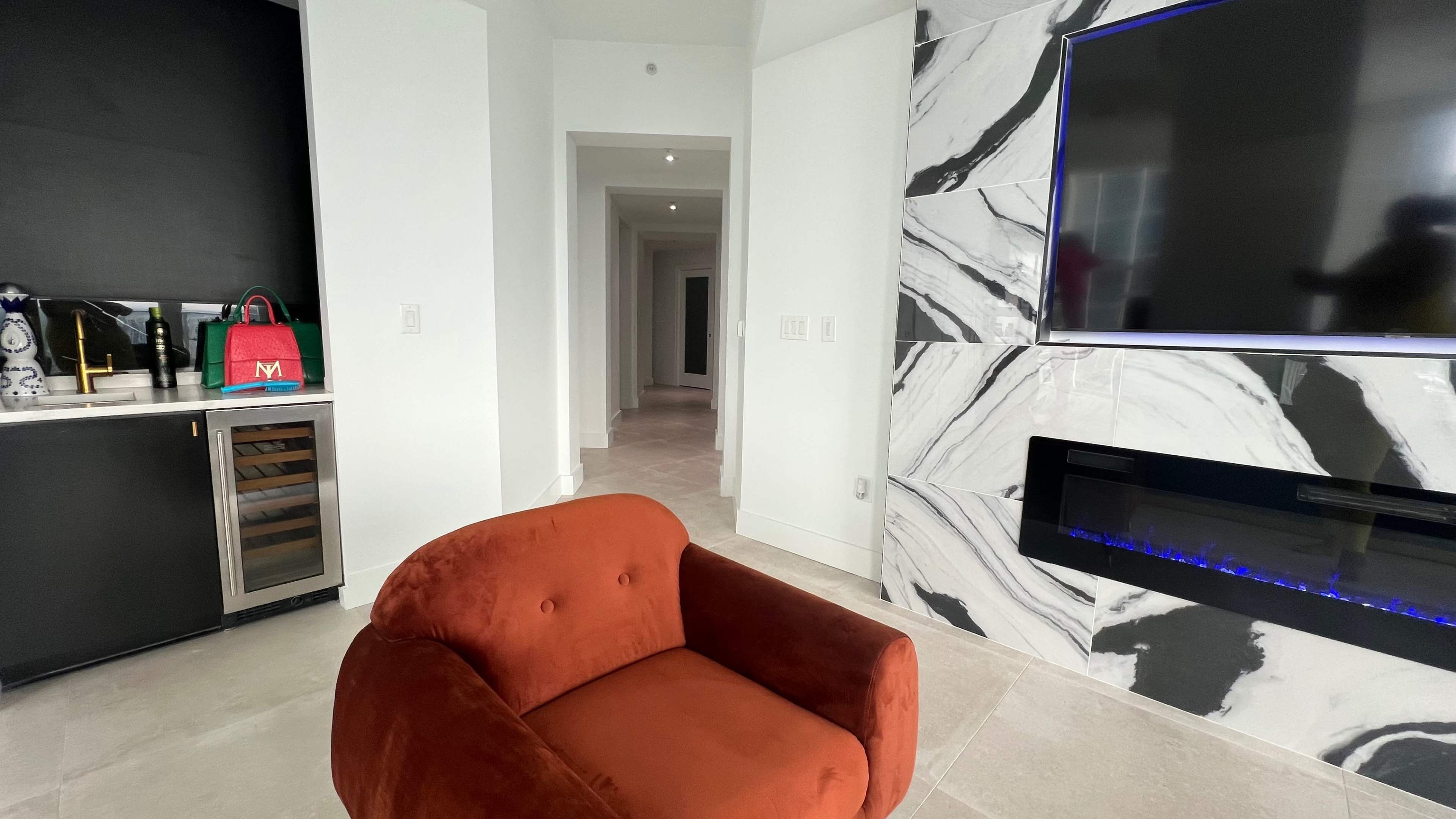 The image shows a modern living space with a brown upholstered chair facing a wall-mounted television, a black and white marble feature wall, and a hallway in the background.