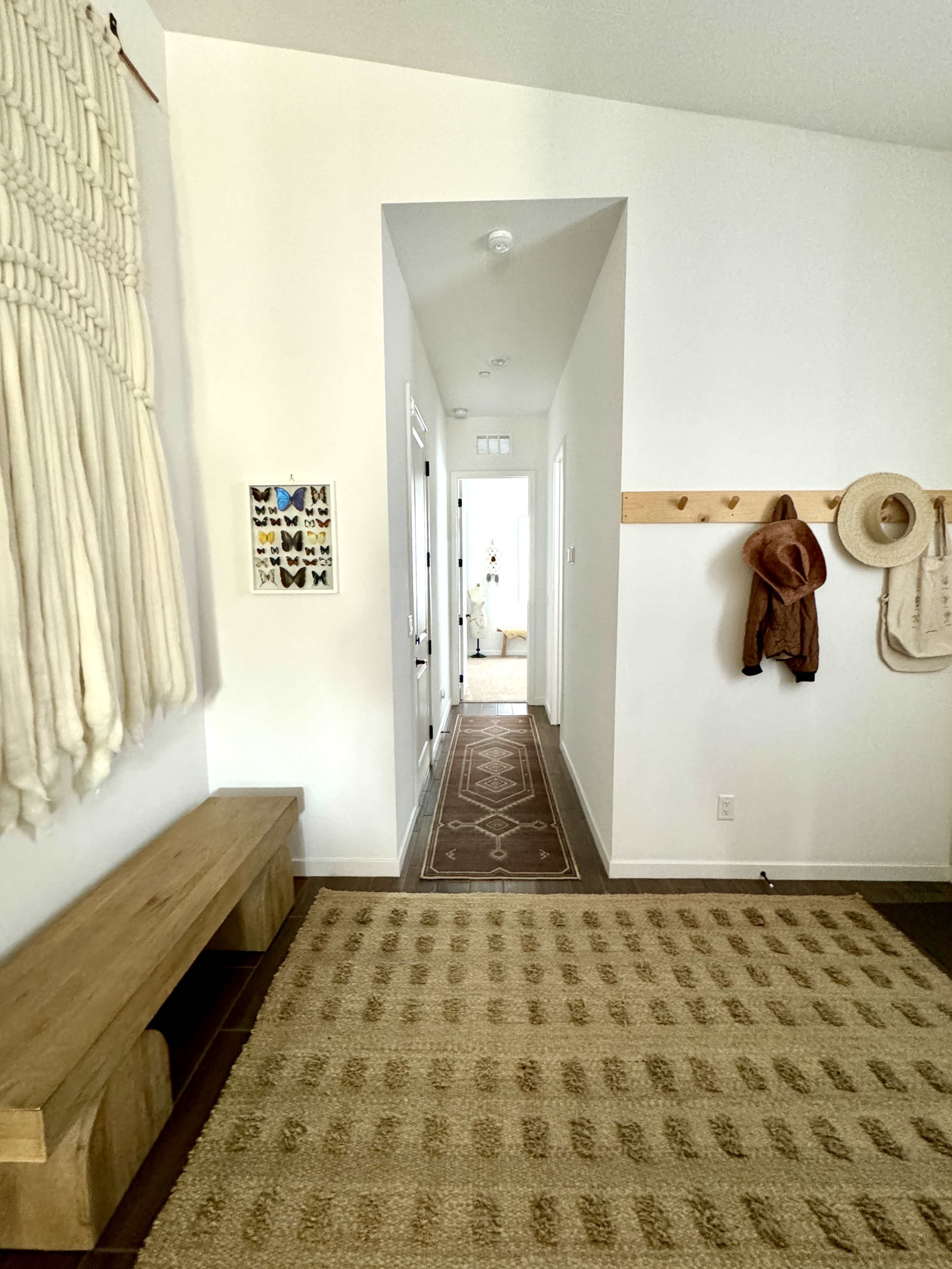 A hallway with a macrame wall hanging, a wooden bench, hooks for coats, and a patterned rug leads to a doorway.