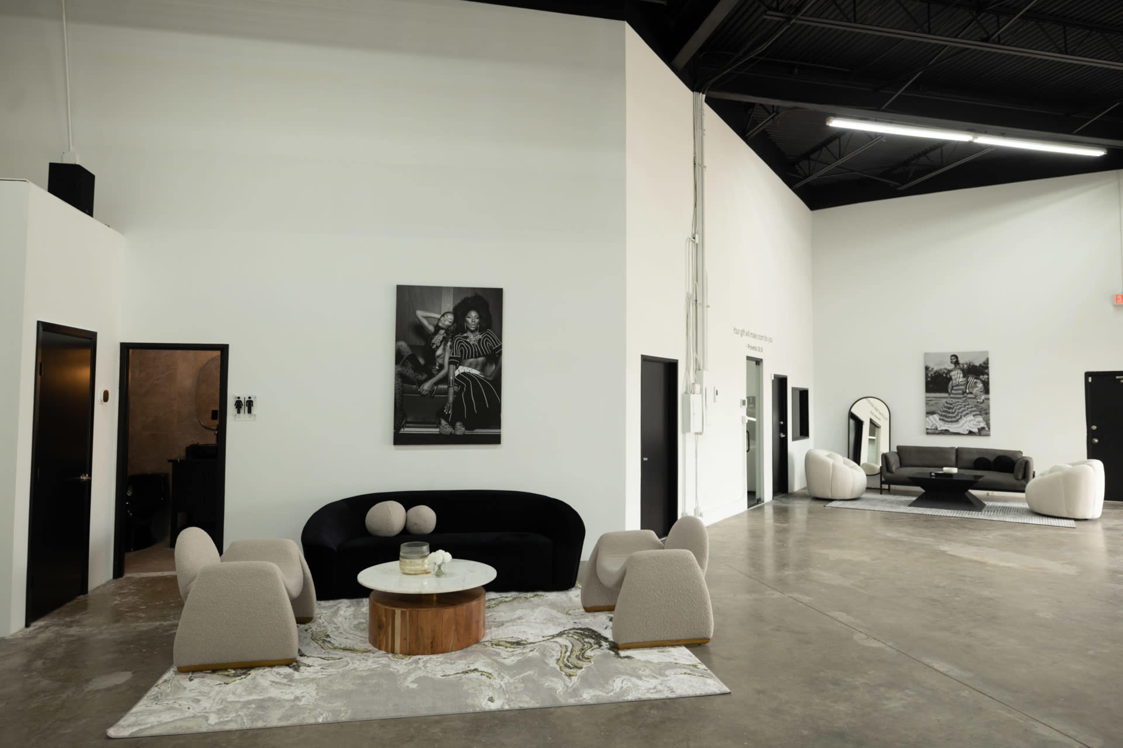 The image shows a modern, spacious interior with minimalist furniture, featuring a black sofa, light-colored chairs, and decorative tables against white walls adorned with black-and-white photographs.