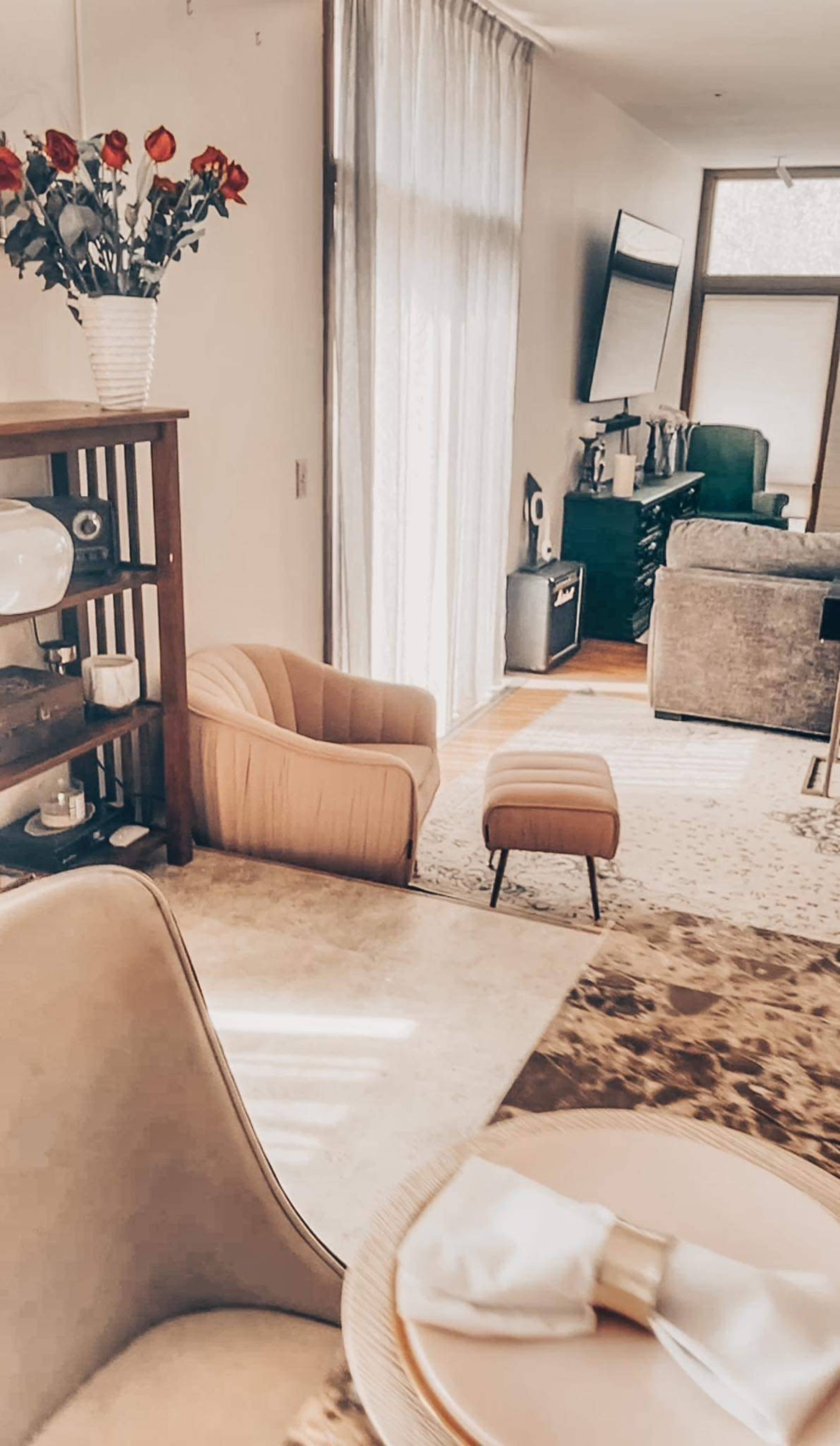 A living room with a beige armchair, an ottoman, and a bookshelf, all illuminated by natural light from large windows.