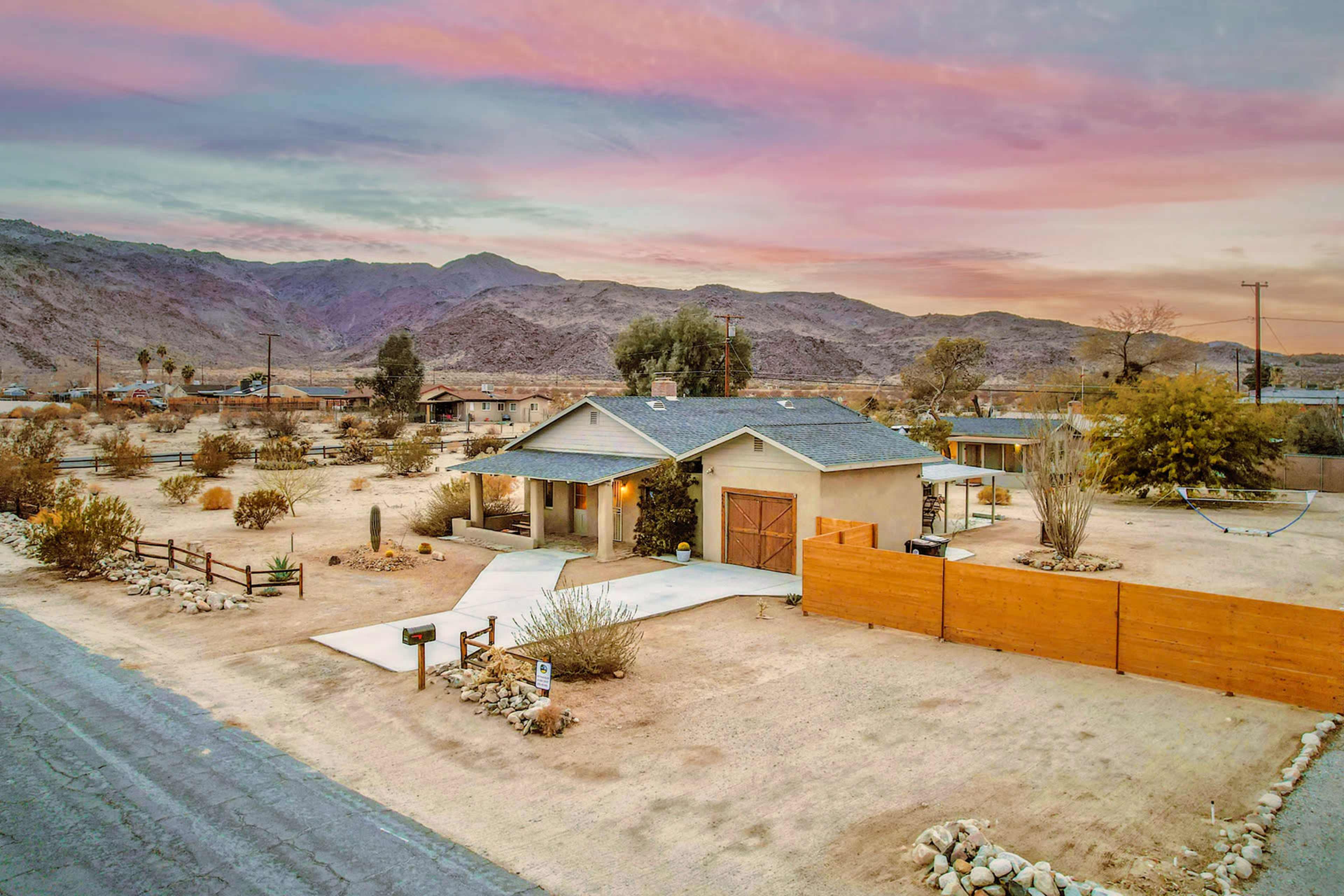 A single-story house sits on a desert landscape surrounded by mountains, with a gravel driveway and wooden fence.