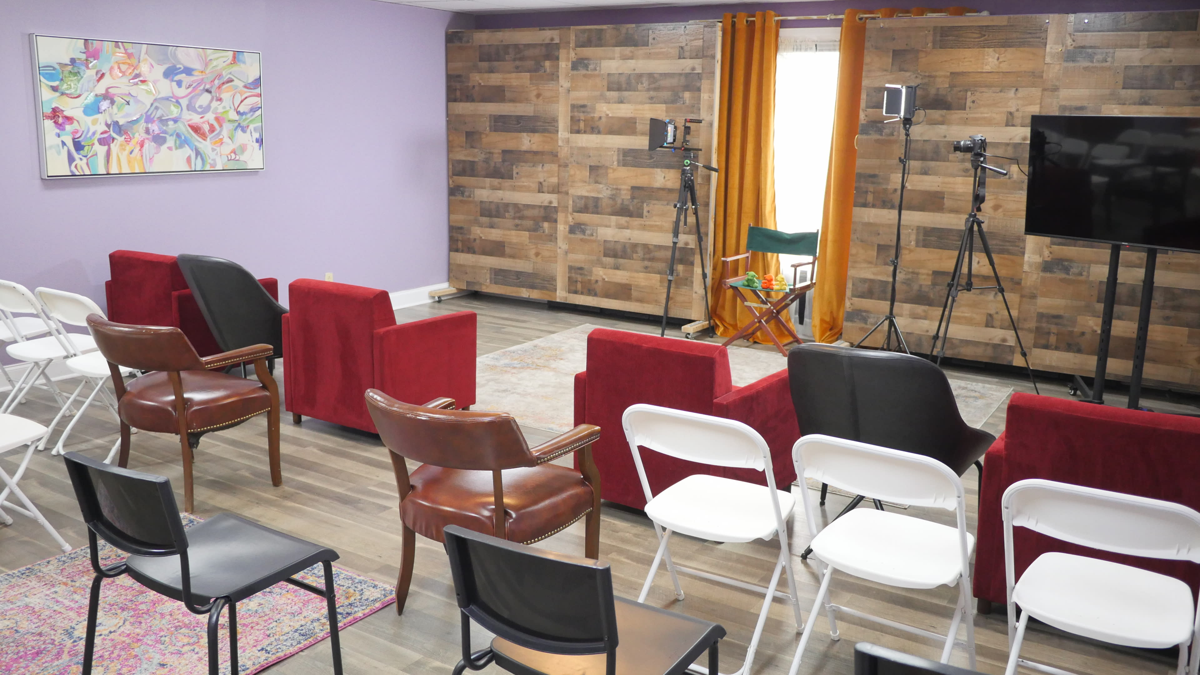 The room features a mix of red and black seating arranged in a semi-circle facing a wooden wall, with two tripods holding cameras and a television stand on one side.