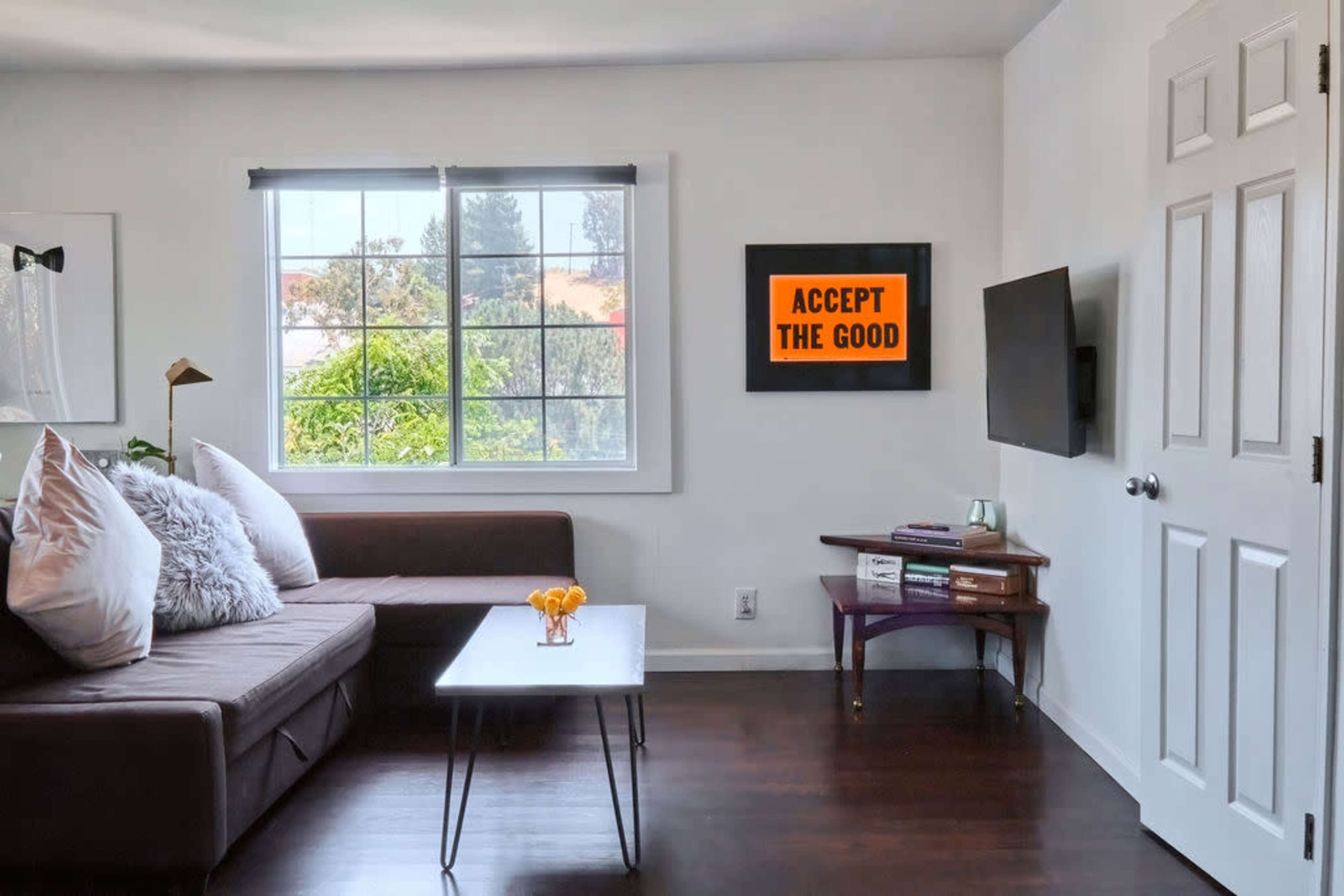 A living room features a brown sectional sofa, a coffee table with a small bowl of oranges, and a framed motivational poster on the wall next to a television.