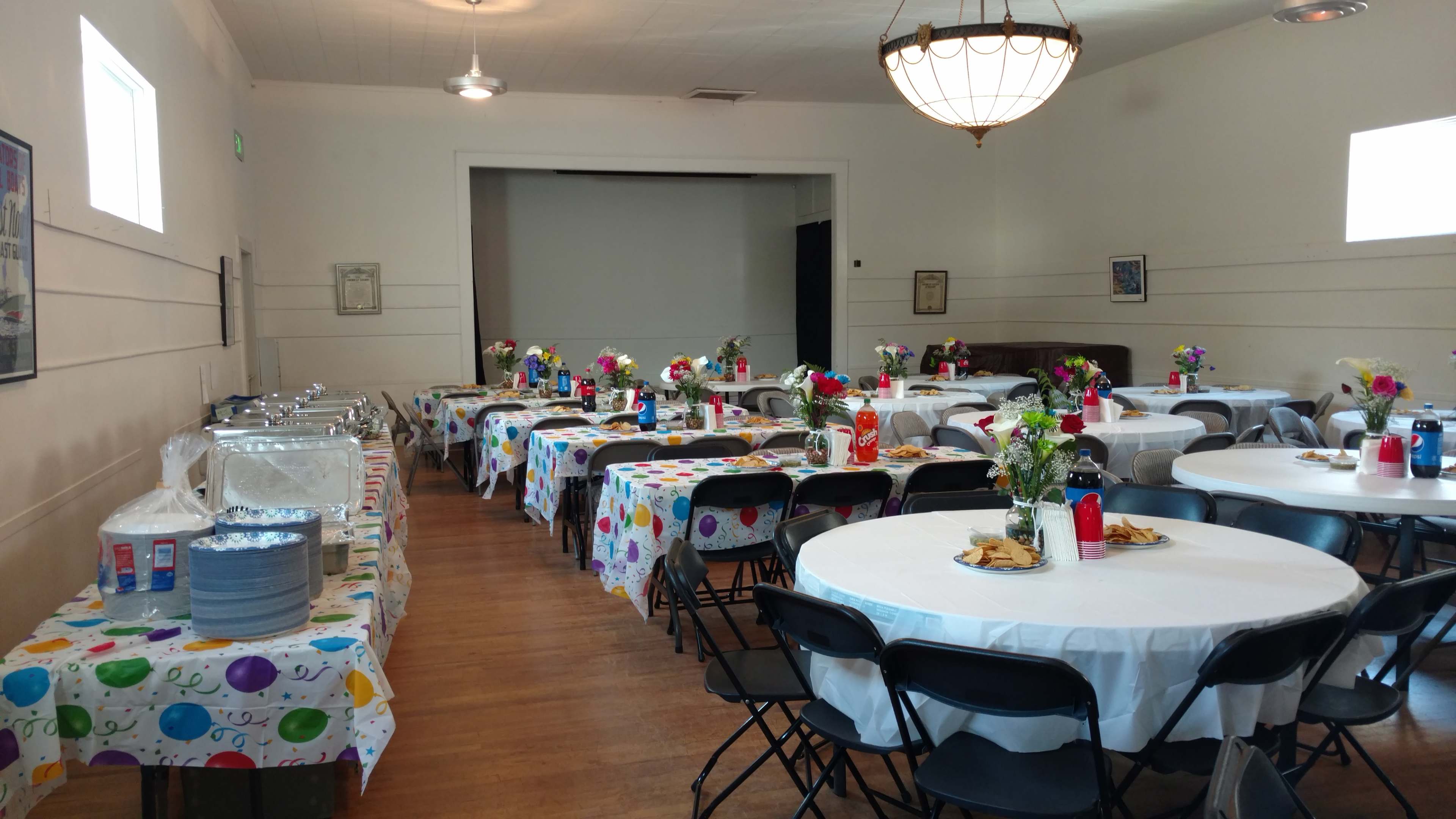 A banquet hall arranged with round tables covered in colorful tablecloths, decorated with flowers, and set up for a gathering.