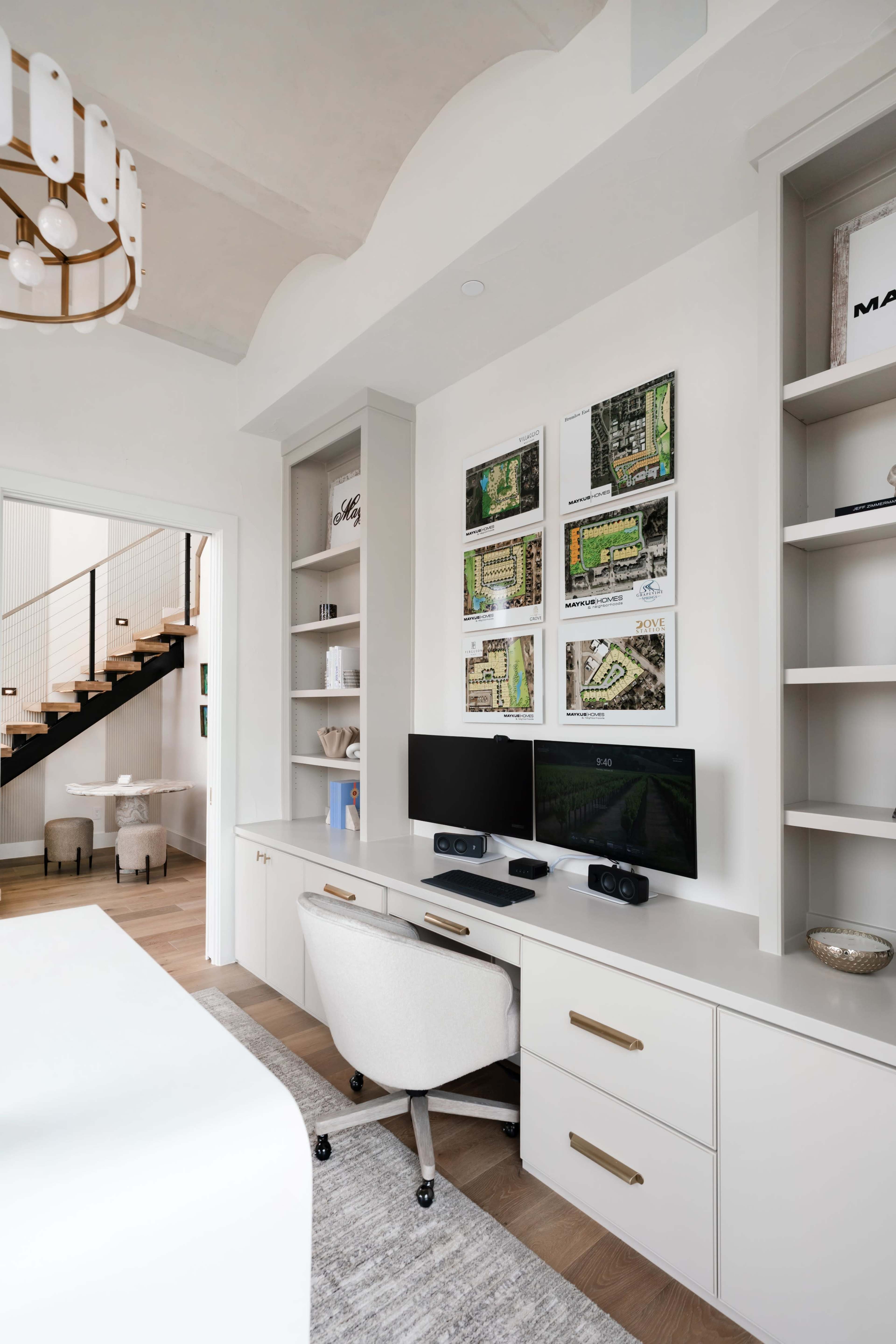 The image shows a modern office space featuring a desk with two monitors, a white swivel chair, and framed maps on the wall, with built-in shelves and a staircase visible in the background.